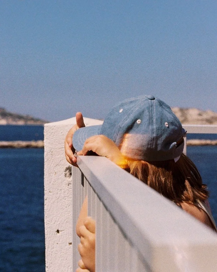 Person wearing a baseball cap and sunglasses leaning over a white fence, with water and land in the background.