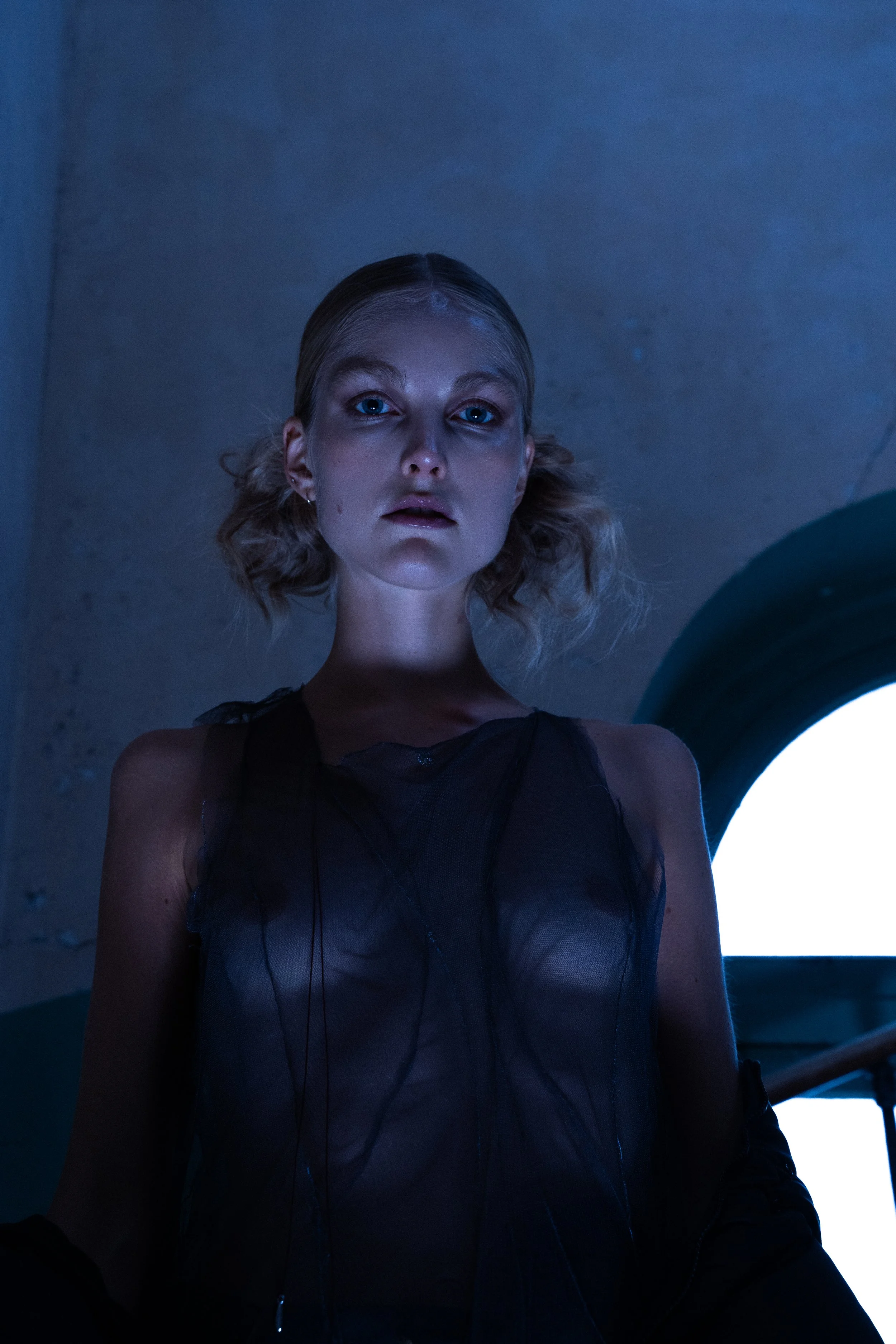 A young woman with blonde hair styled in loose curls, wearing a sheer black top, stands in dim lighting with a blue hue, with a bright window in the background.
