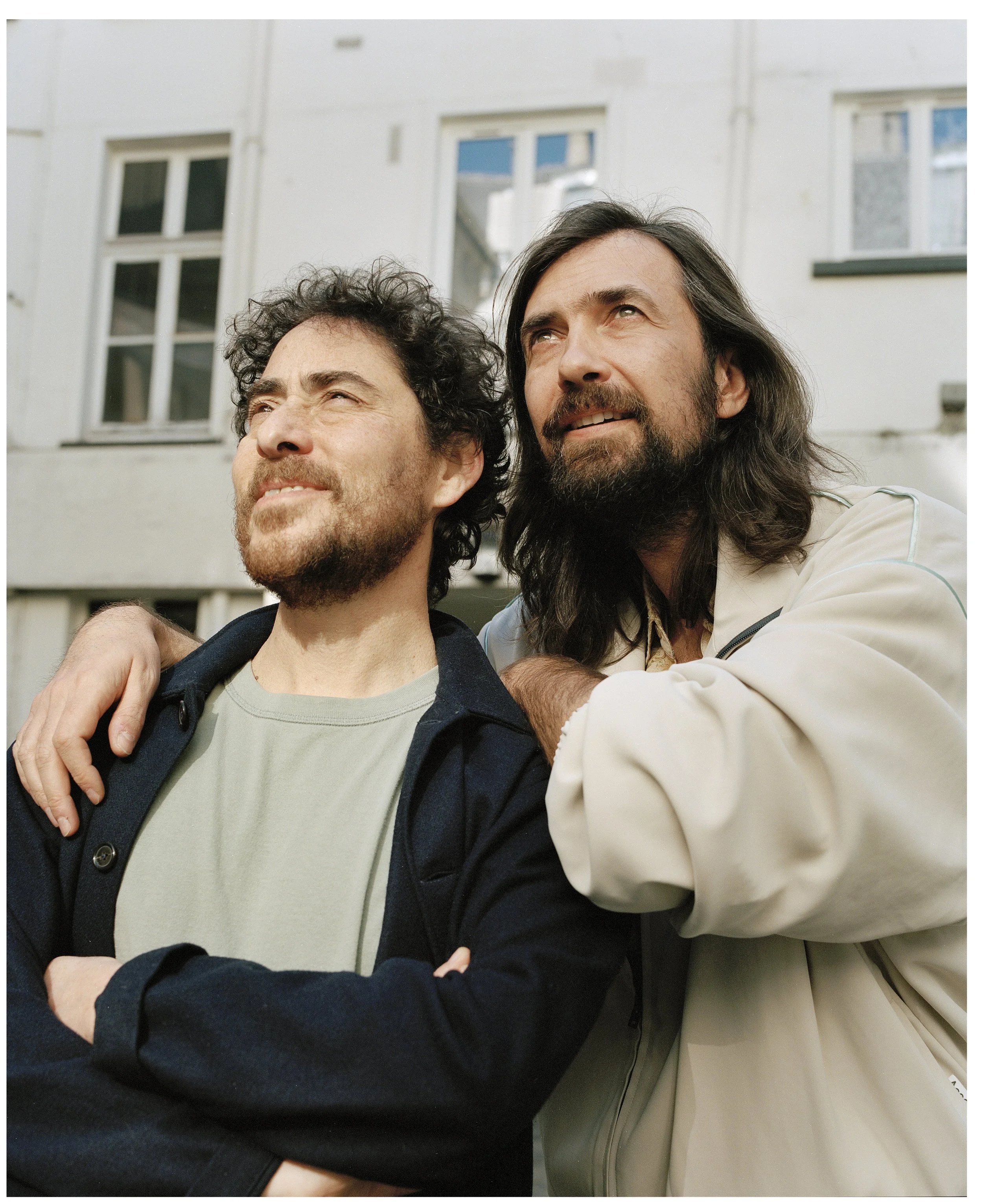 Two men standing close together outdoors, one with his arm around the other's shoulder, looking off into the distance with a smile, against a background of a light-colored building with several windows.