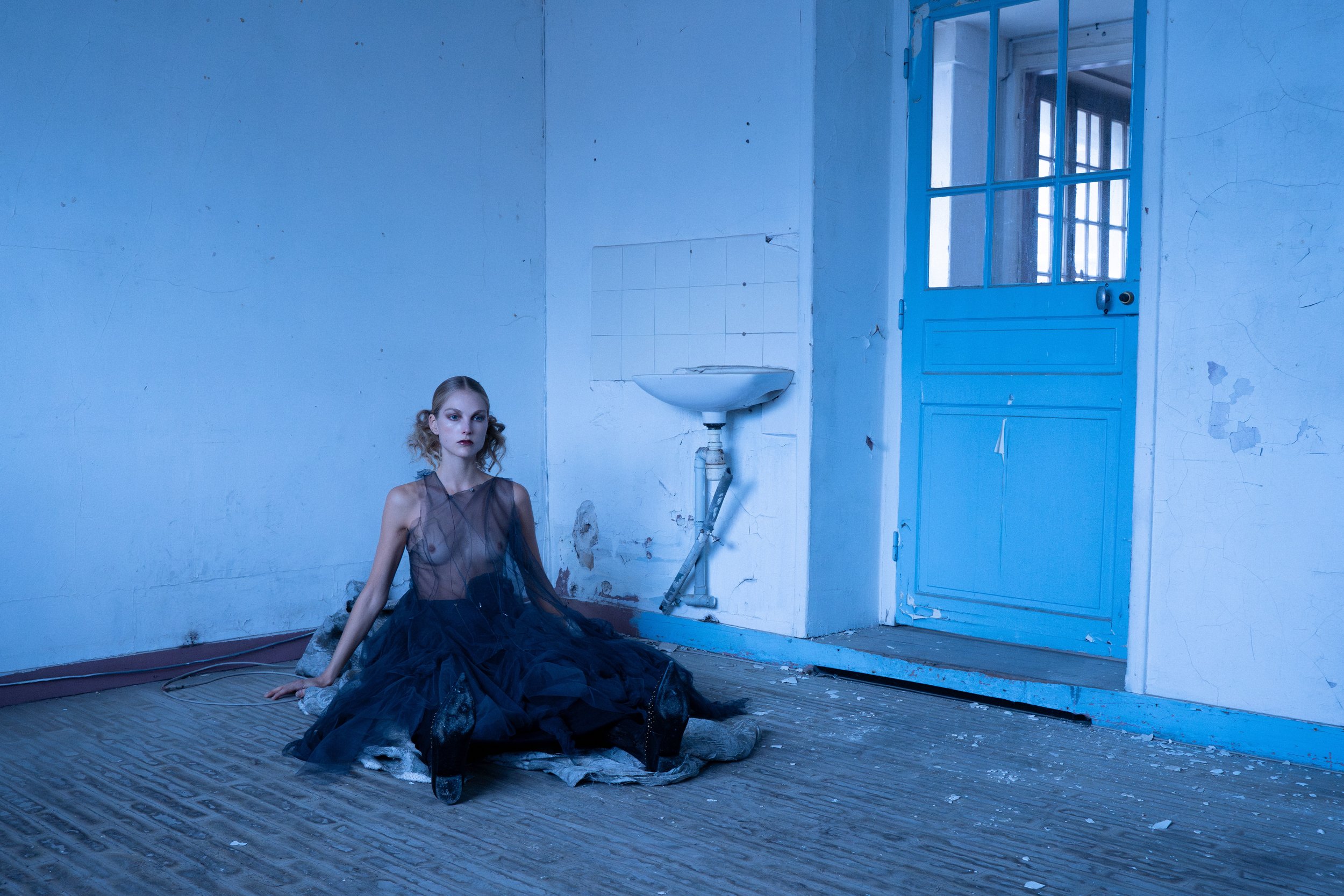 A woman with blonde hair, dressed in a sheer black top and a dark skirt, sits on the floor in a dilapidated room with peeling paint and a blue door. The room appears abandoned and run-down.