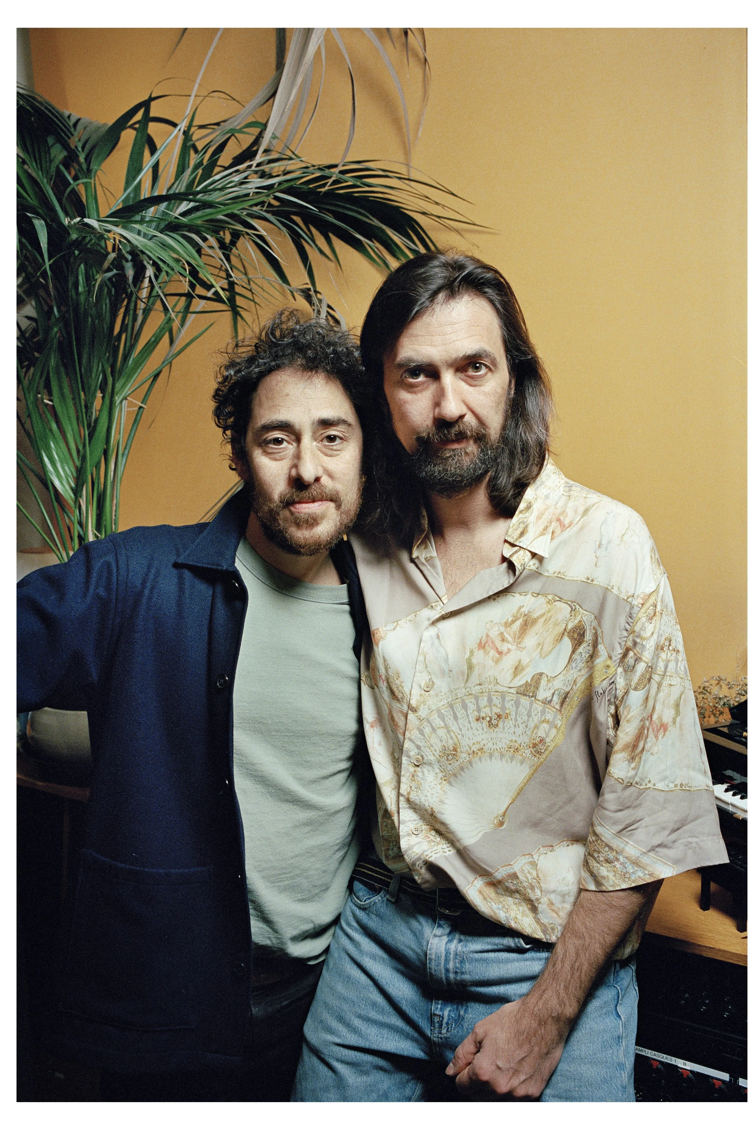 Two men with dark hair and beards standing close to each other, one wearing a light-colored patterned shirt and the other in a dark jacket and light shirt, in front of a yellow wall with a large green plant behind them.