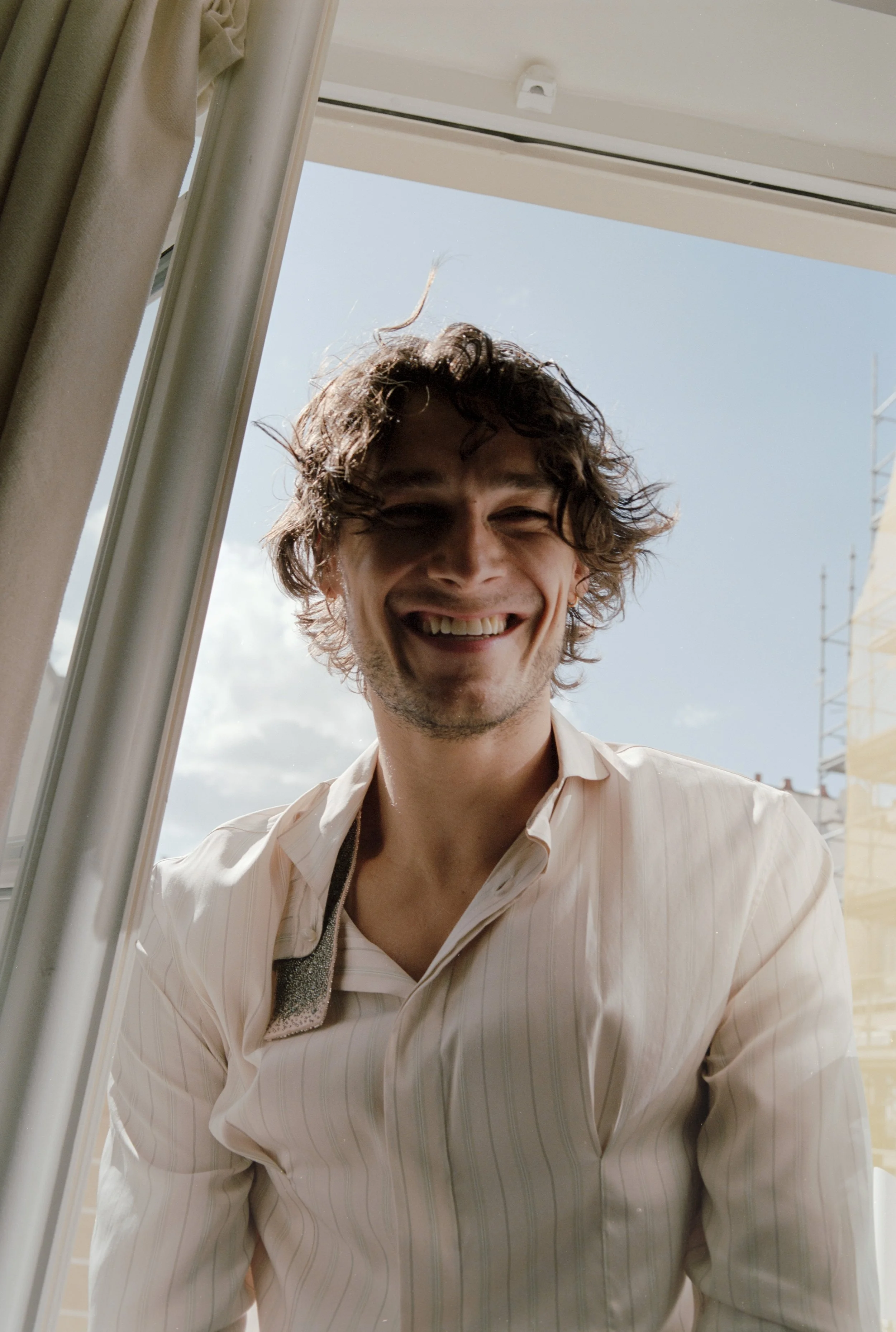 A young man with curly brown hair smiling and looking at the camera, standing by a window with a partly cloudy sky in the background.