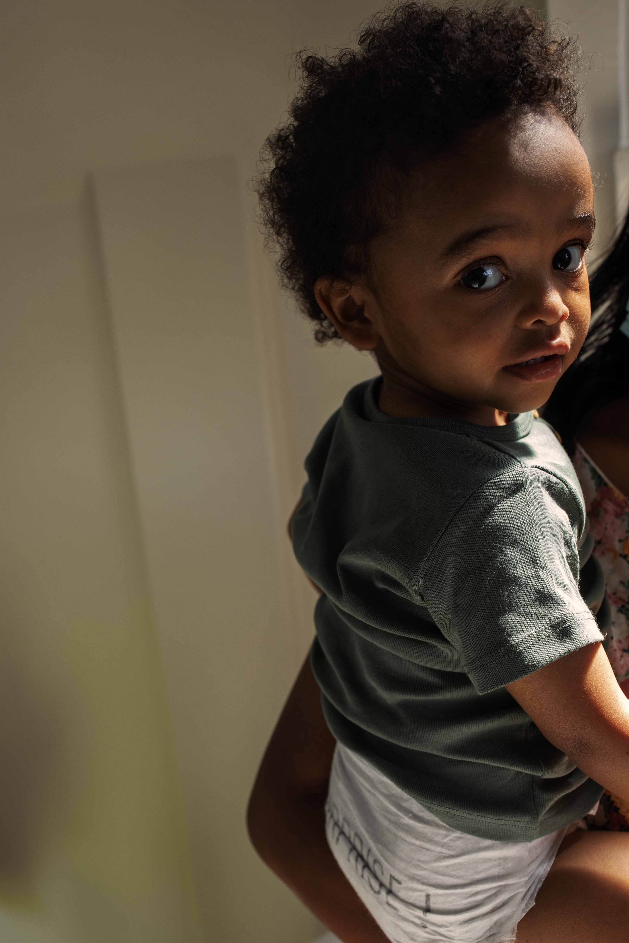 Close-up of a young child with curly hair, wearing a green t-shirt and white shorts, being held by an adult. The child is looking at the camera with a slightly open mouth and wide eyes.