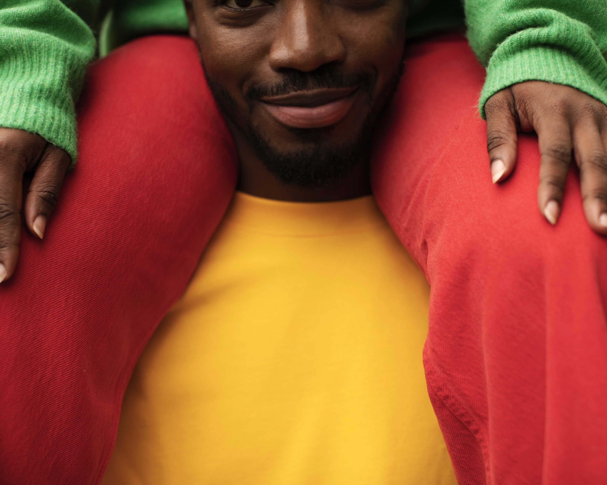 A man smiling with a child on his shoulders, wearing a yellow shirt, red pants, and a green sweater.