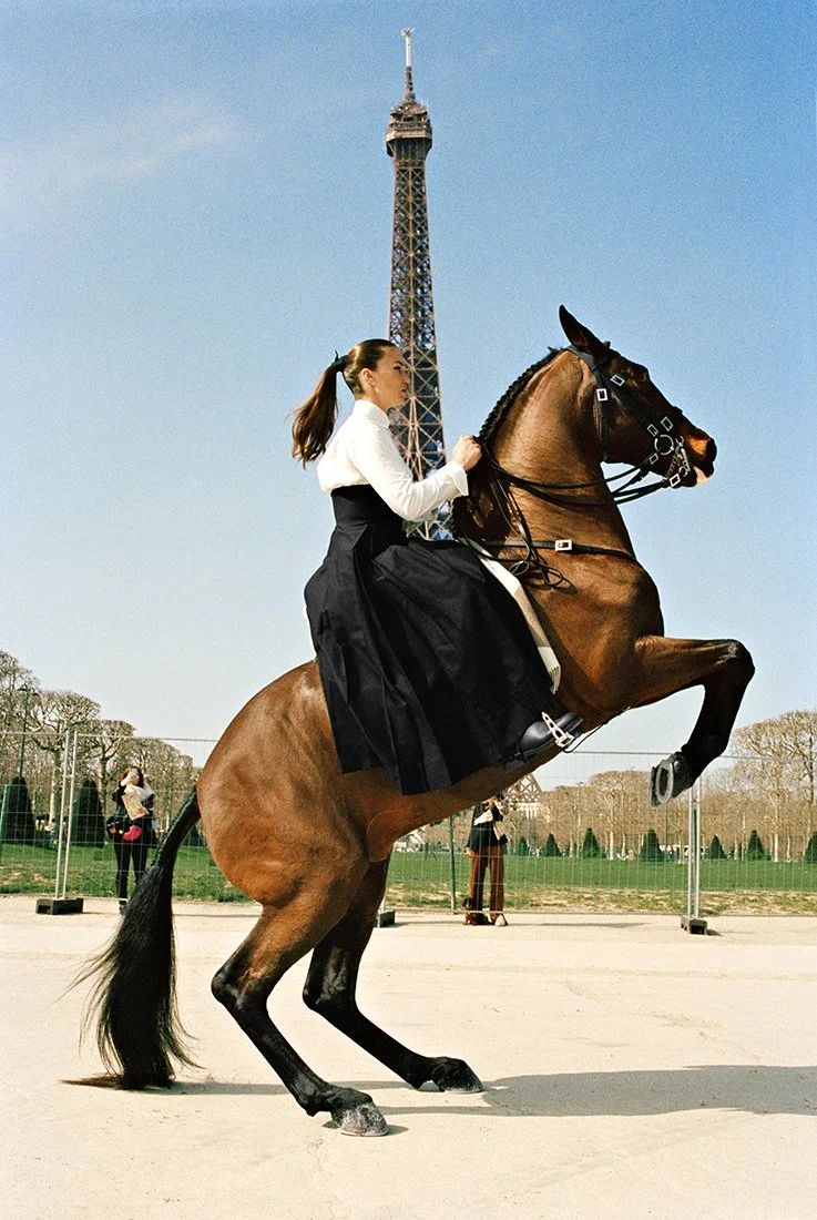 A woman riding a rearing horse in front of the Eiffel Tower during the day, with other people and a fence in the background.