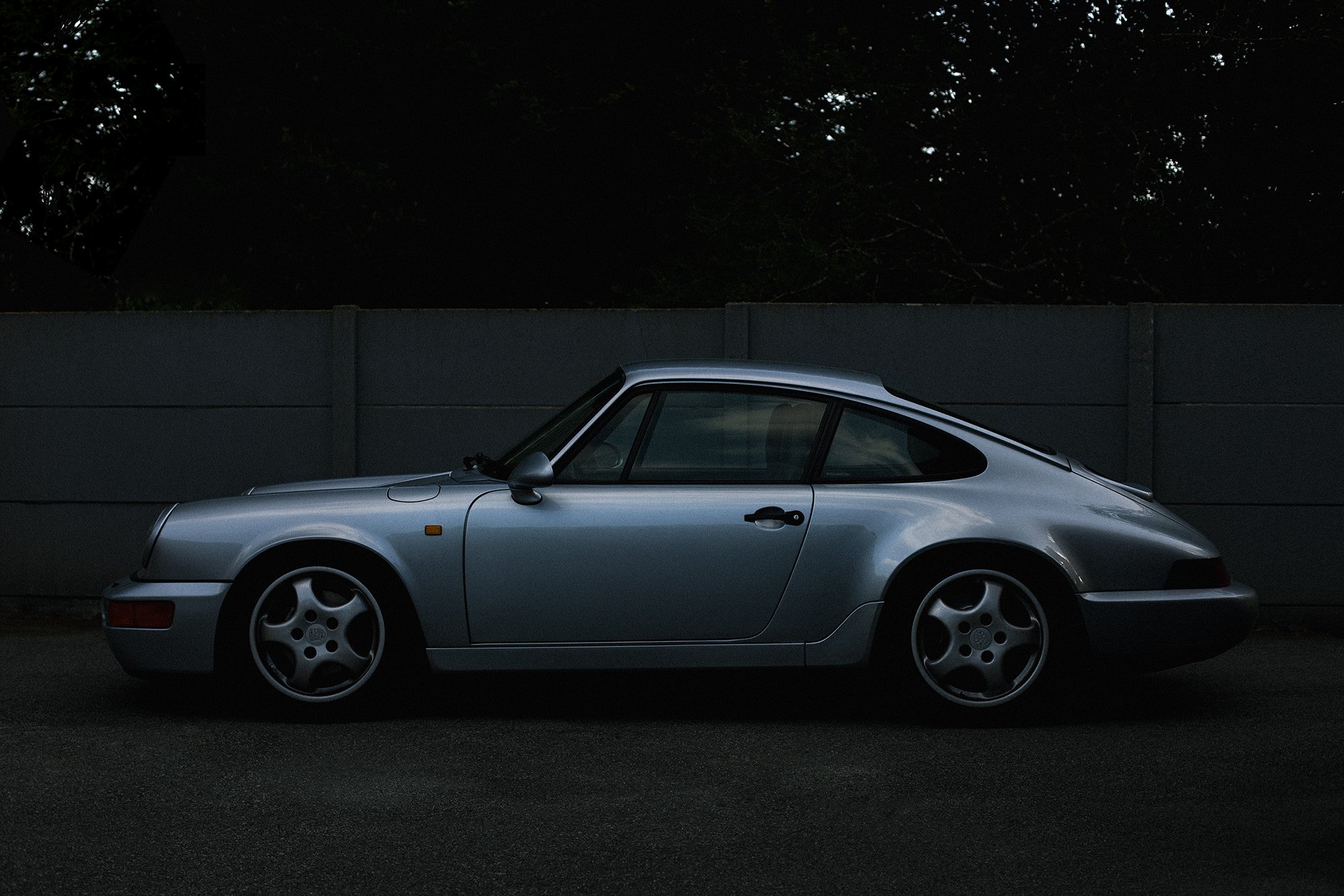 A classic silver Porsche 911 sports car parked on a dark street with a dark fence and trees in the background.
