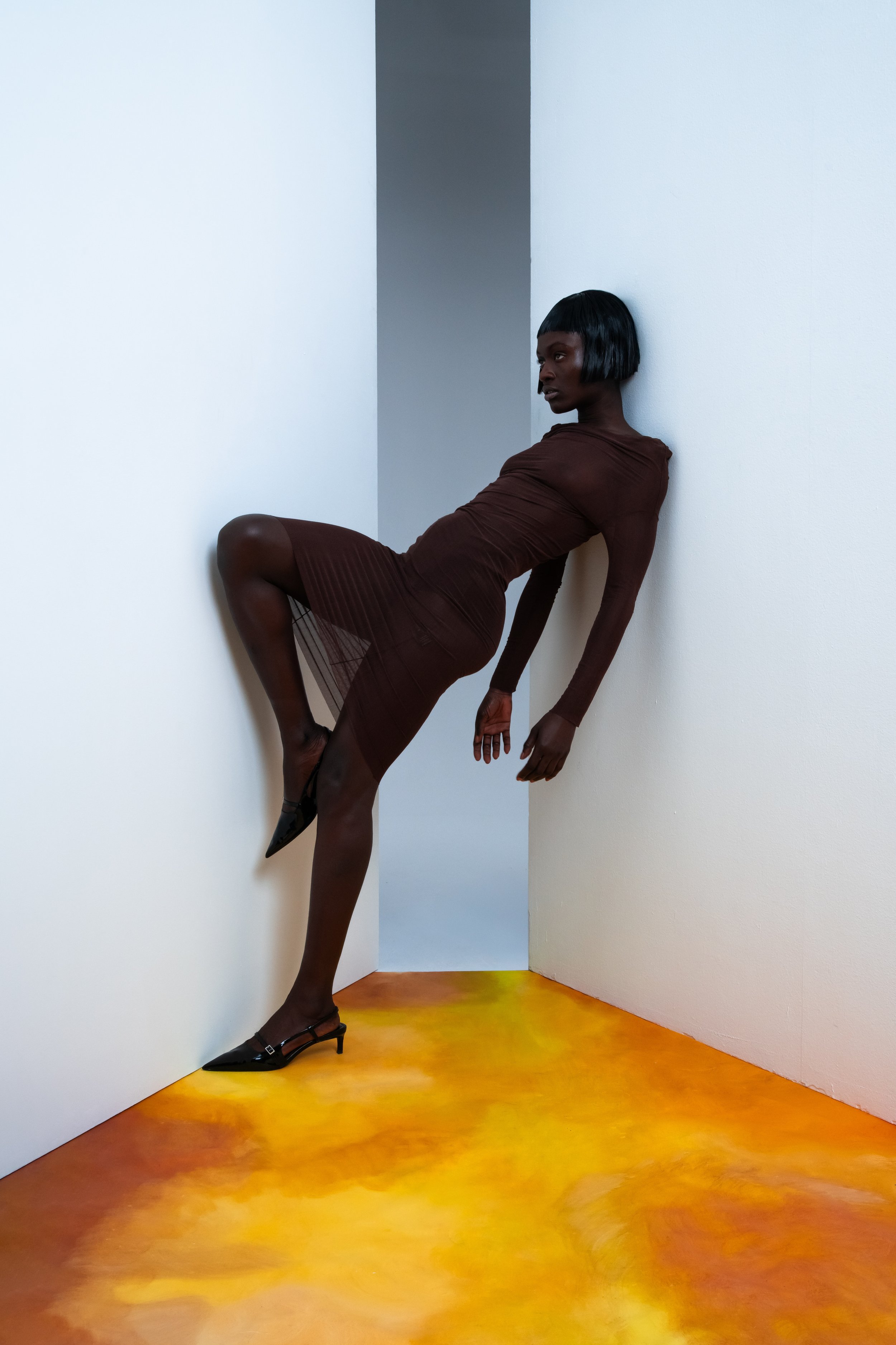 A woman with dark skin and a bob haircut wearing a brown dress and heels, posing with her back against a white wall in a modern, minimalistic space with a yellow-orange floor.