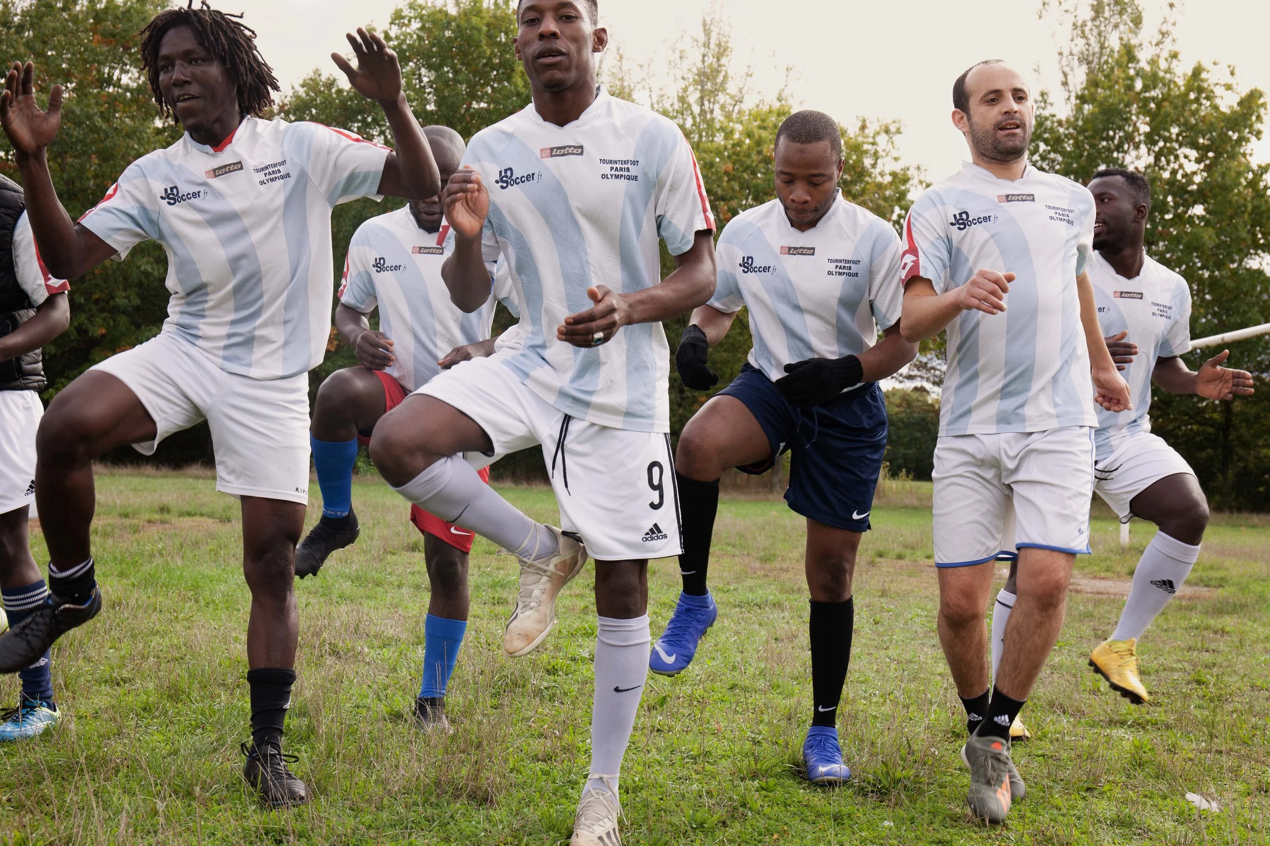 Group of diverse men in soccer uniforms doing warm-up exercises on a grassy field outdoors.