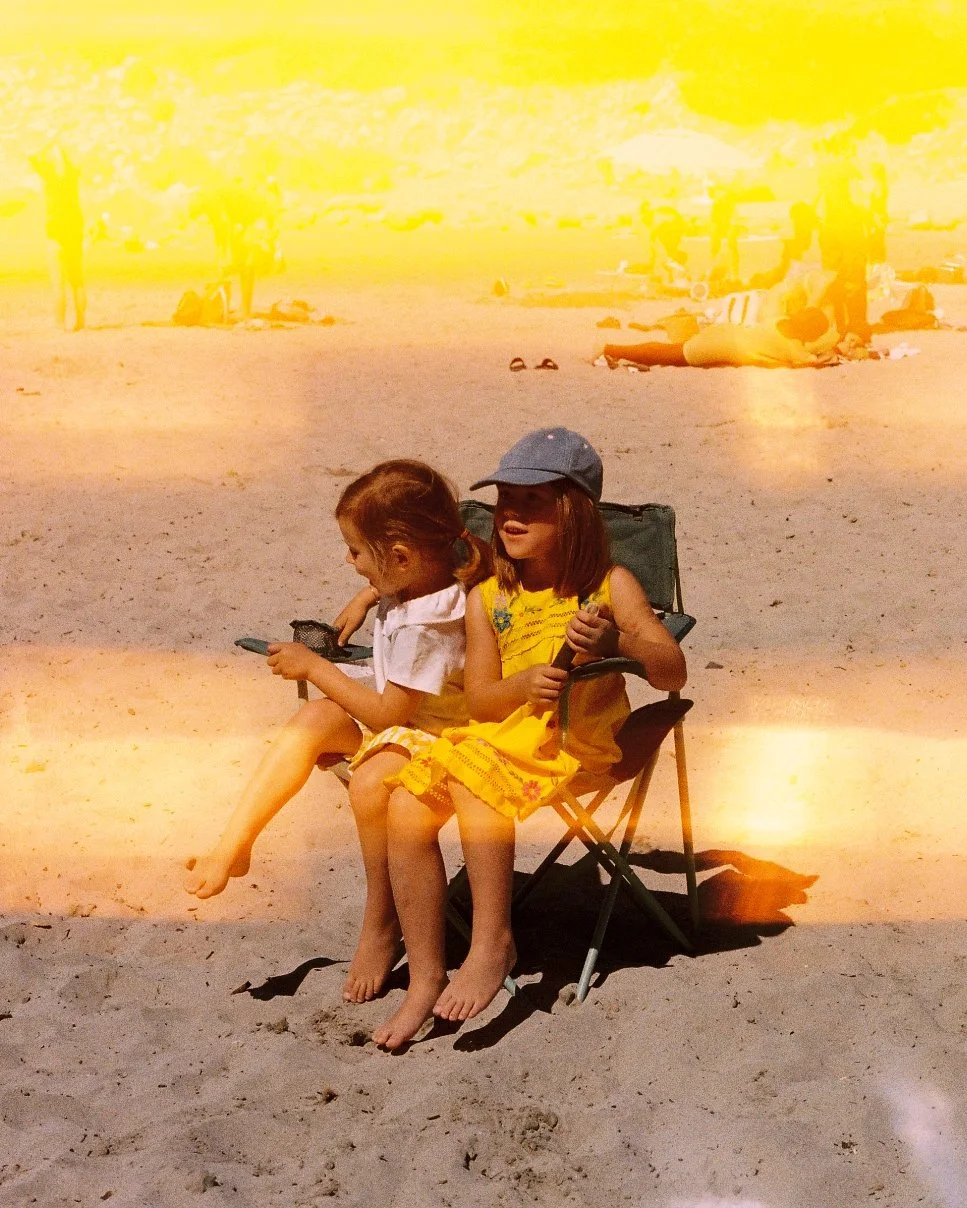 Two young girls sitting on a beach chair, one wearing a yellow dress and gray cap, the other in a white shirt, with people relaxing and walking on the beach in the background.