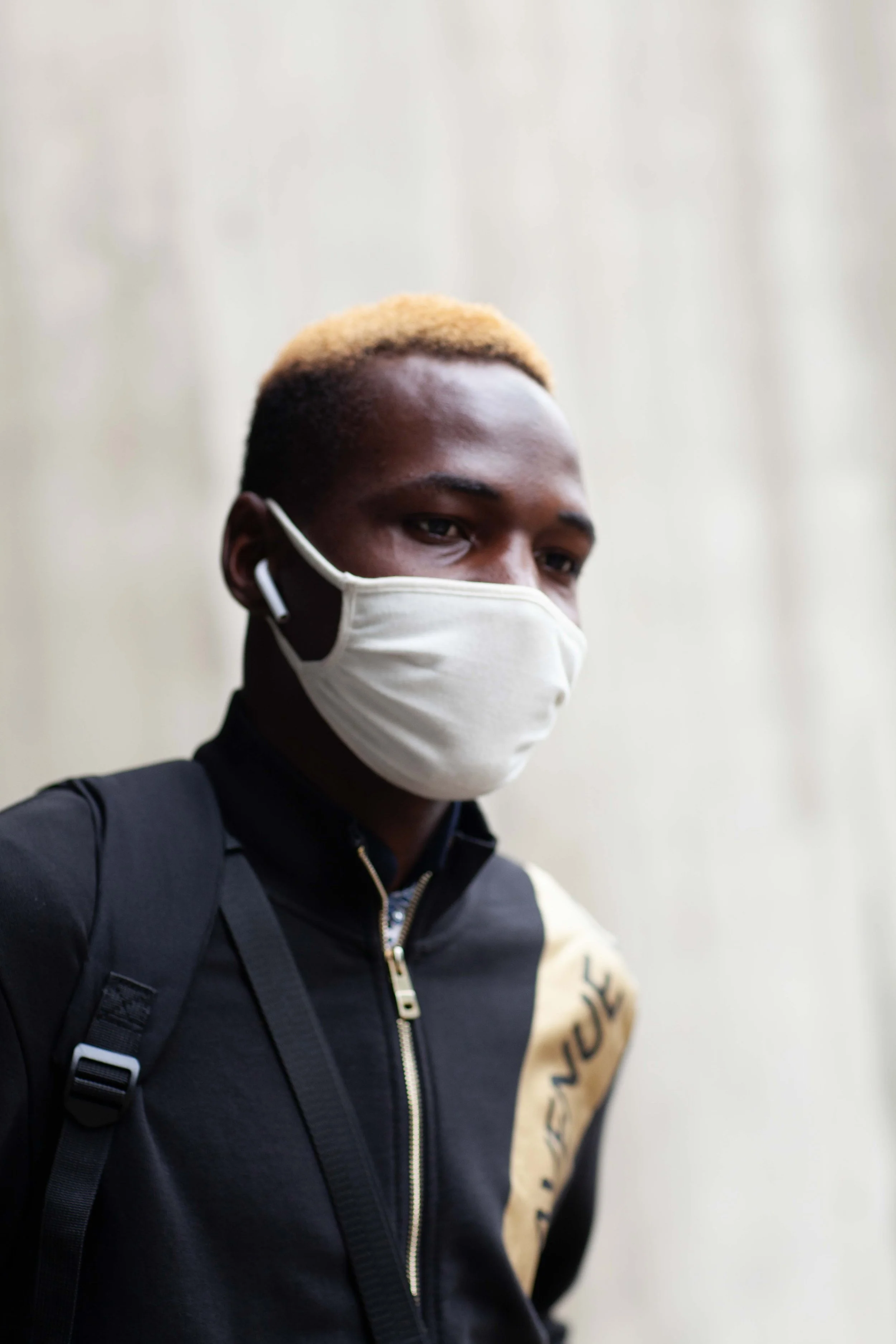 A young man wearing a white face mask, black hoodie with a beige and black design, and a backpack, standing against a light-colored wall.