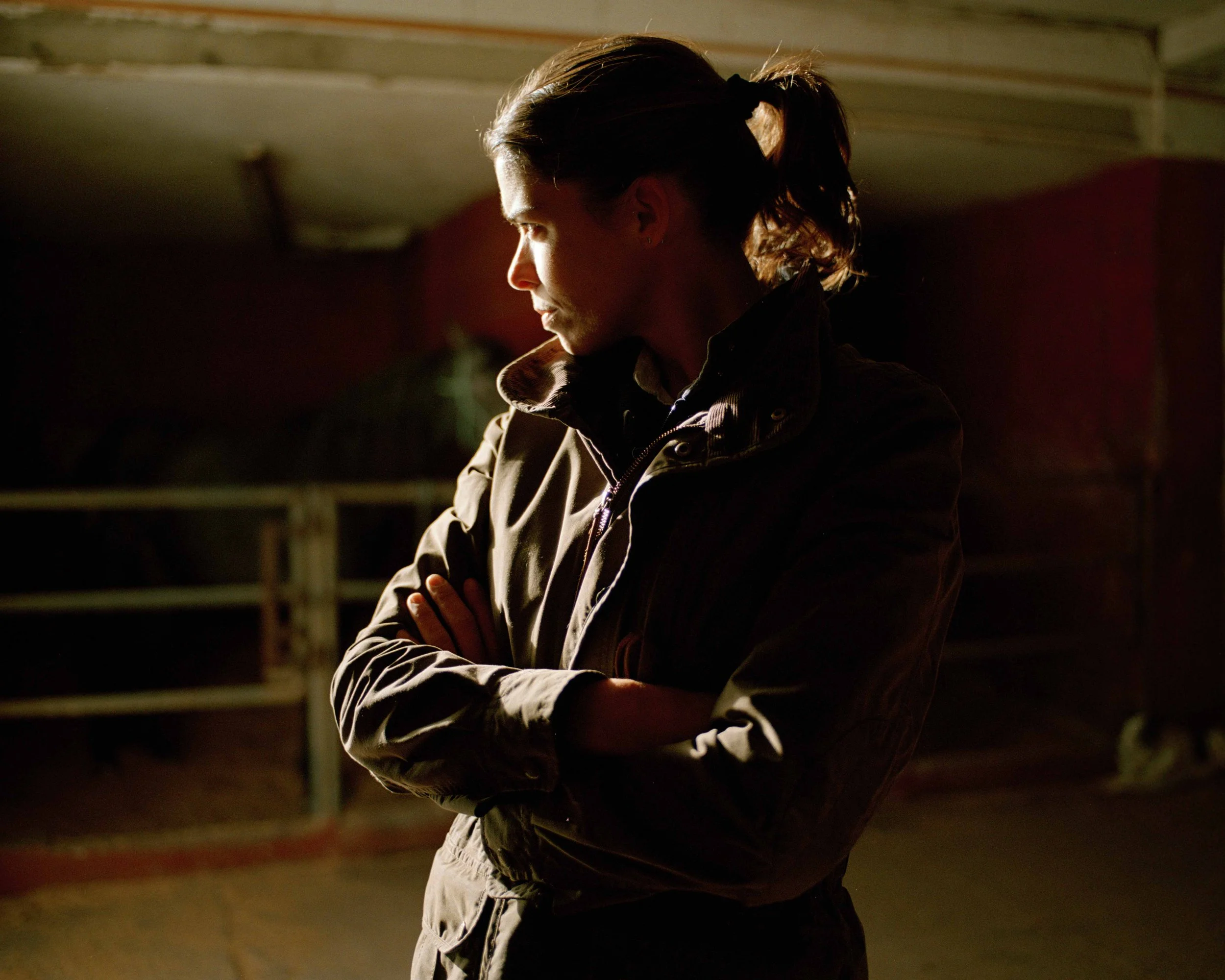 Side profile of a woman with arms crossed, wearing a jacket, in a dimly lit setting.