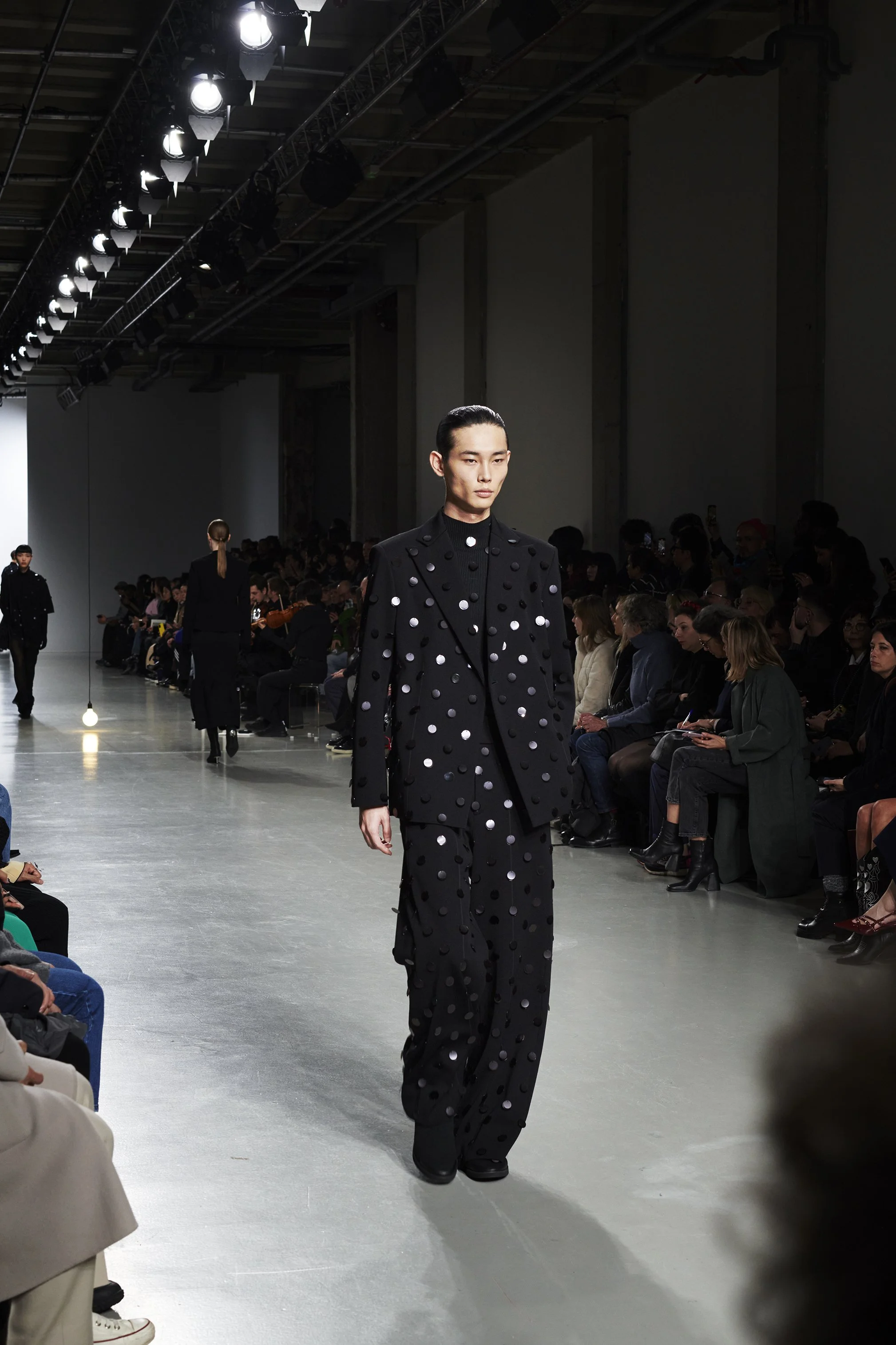 Model walking the runway in a black suit with white polka dots during a fashion show with audience seated on both sides.