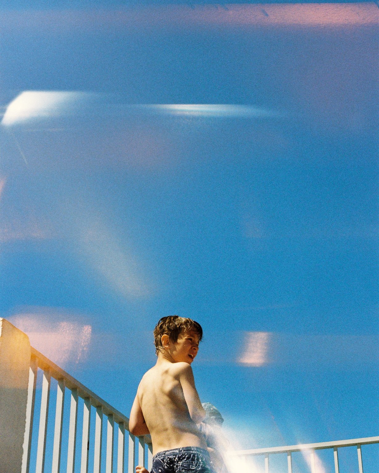 A shirtless boy with dark hair, wearing dark swim trunks, standing on a white poolside railing, looking over his shoulder against a bright blue sky.