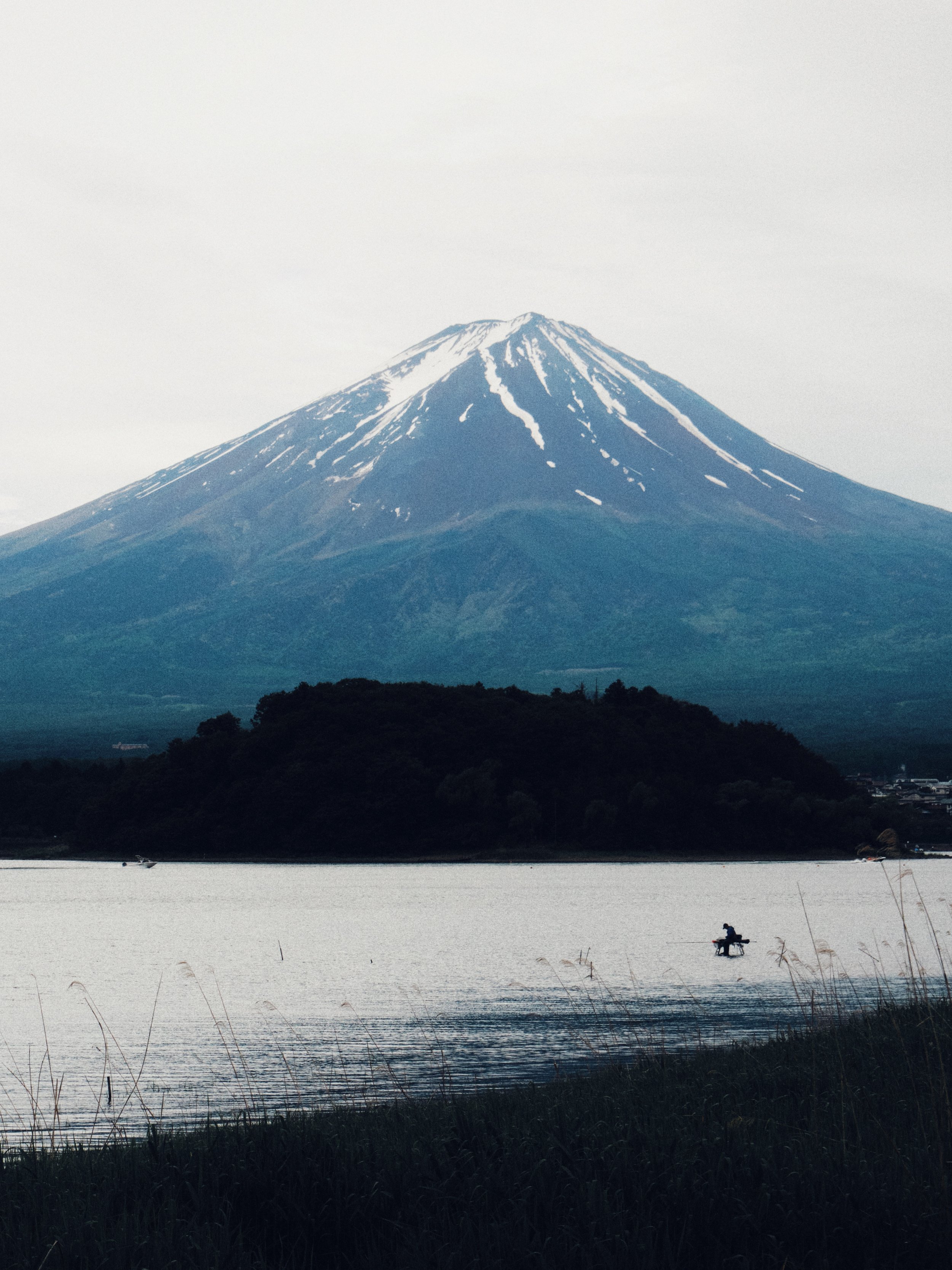 Mount Fuji with snow on its peak, seen across a body of water with a person fishing in a small boat and some grasses in the foreground.