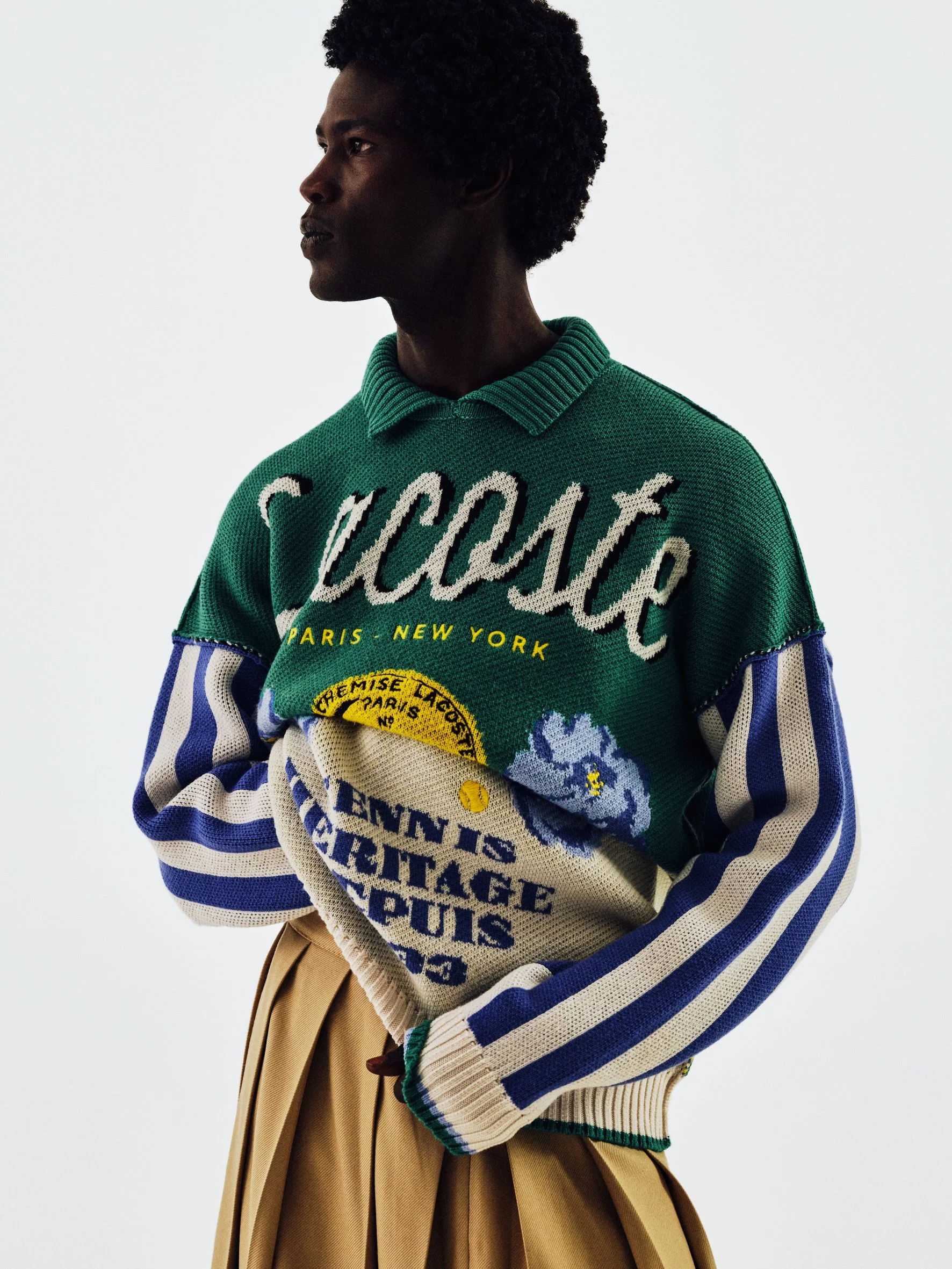 Person wearing a colorful, patterned sweater with various words and designs, beige pleated skirt, standing against a plain light background.