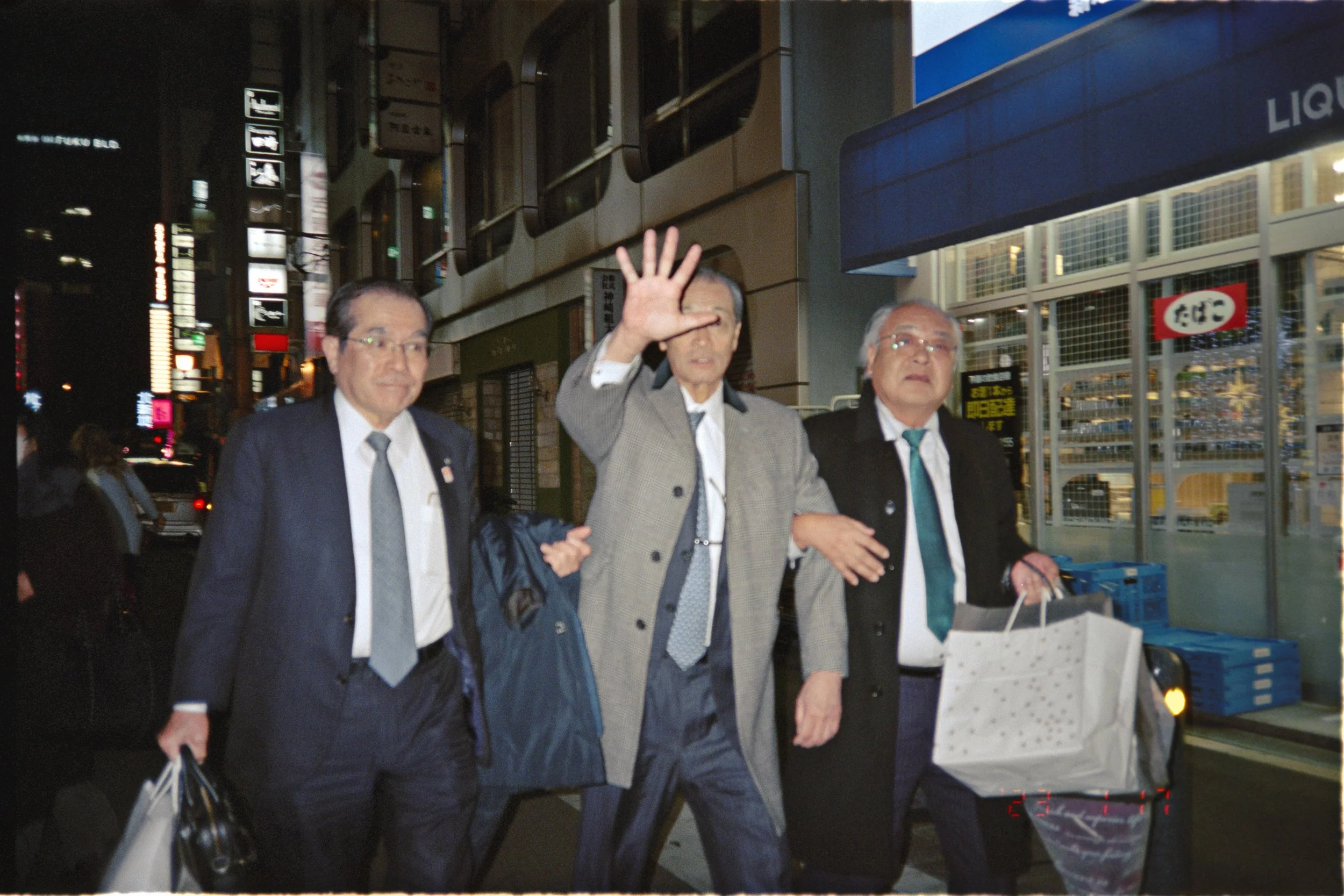 Three men in suits walking on a city street at night, with the man in the center waving, carrying shopping bags.