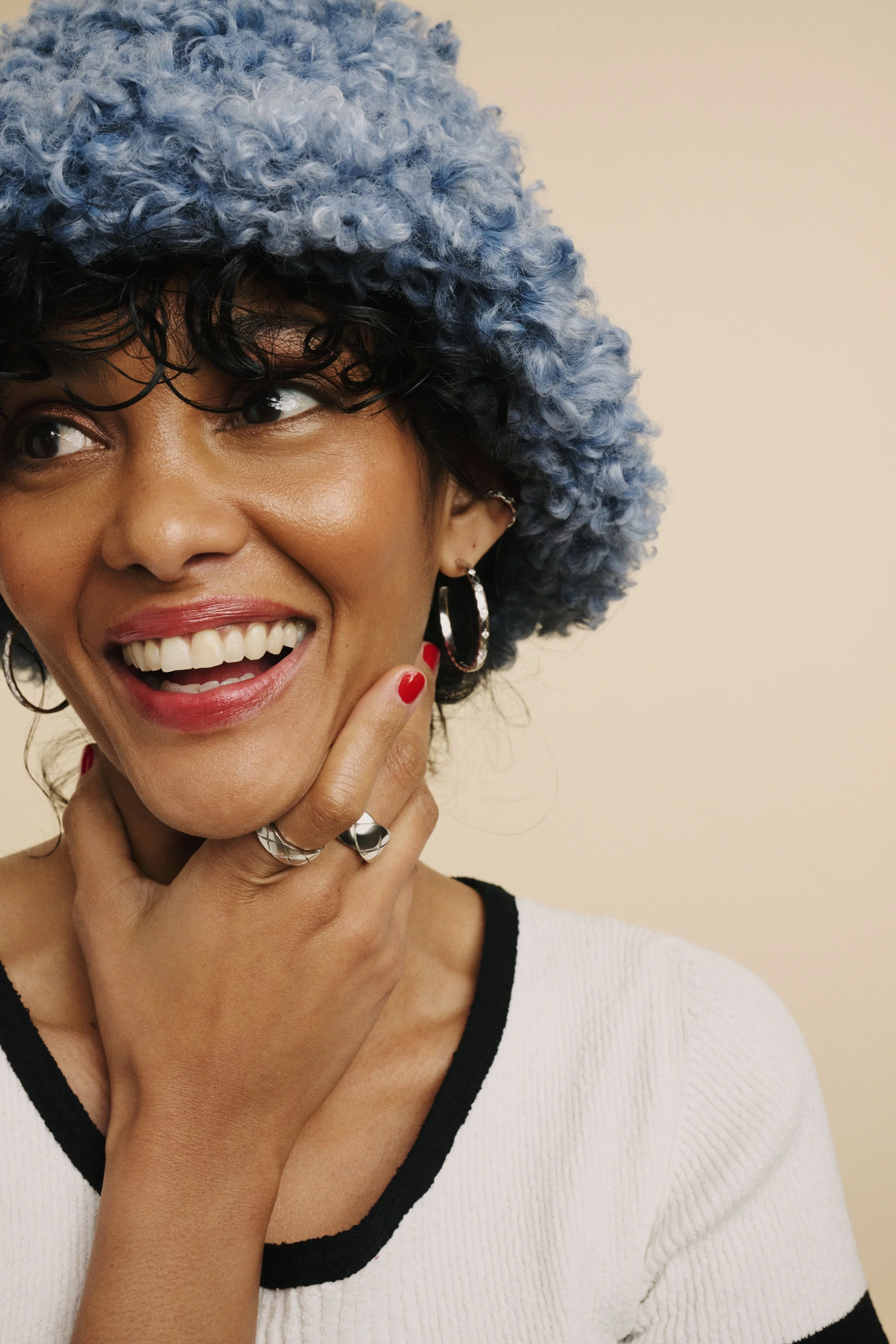 A woman smiling and touching her chin, wearing a fuzzy blue hat, hoop earrings, and rings, with red nail polish, in front of a plain beige background.