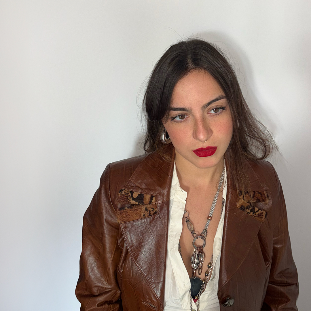 A woman with dark hair, red lipstick, wearing a brown leather jacket with animal print patches, layered necklaces, and hoop earrings, standing against a plain white wall.