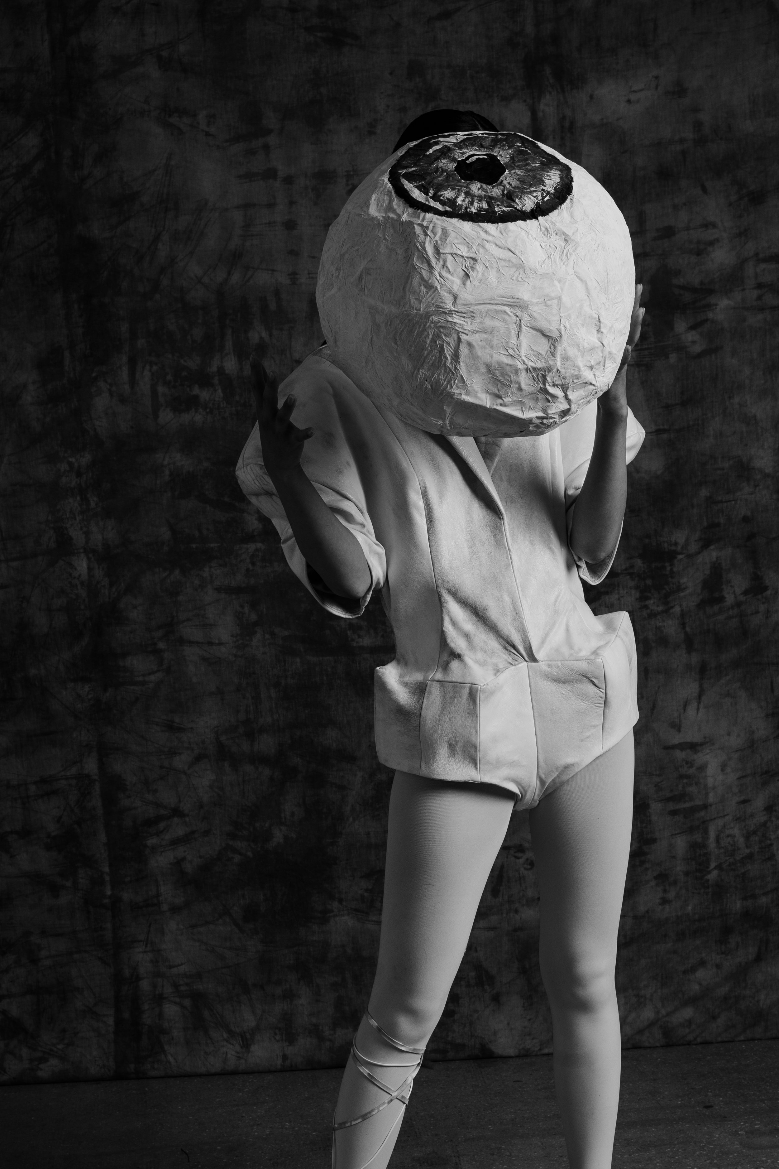 Person wearing a costume with a large paper-mâché head resembling an eye, posing against a textured dark background.