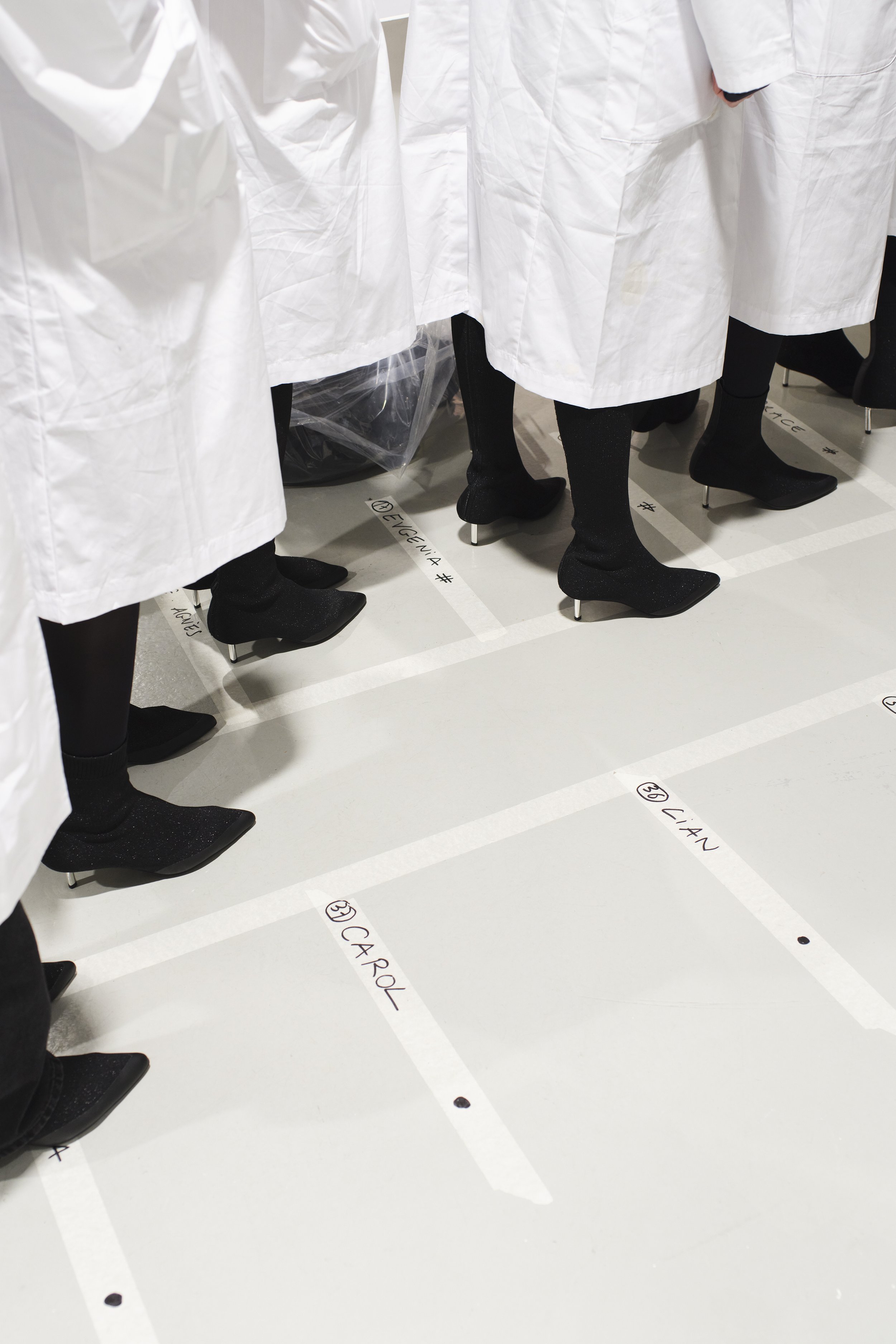 Group of people wearing white lab coats and black high-heeled boots standing on marked grid lines on the floor in a laboratory or medical setting.