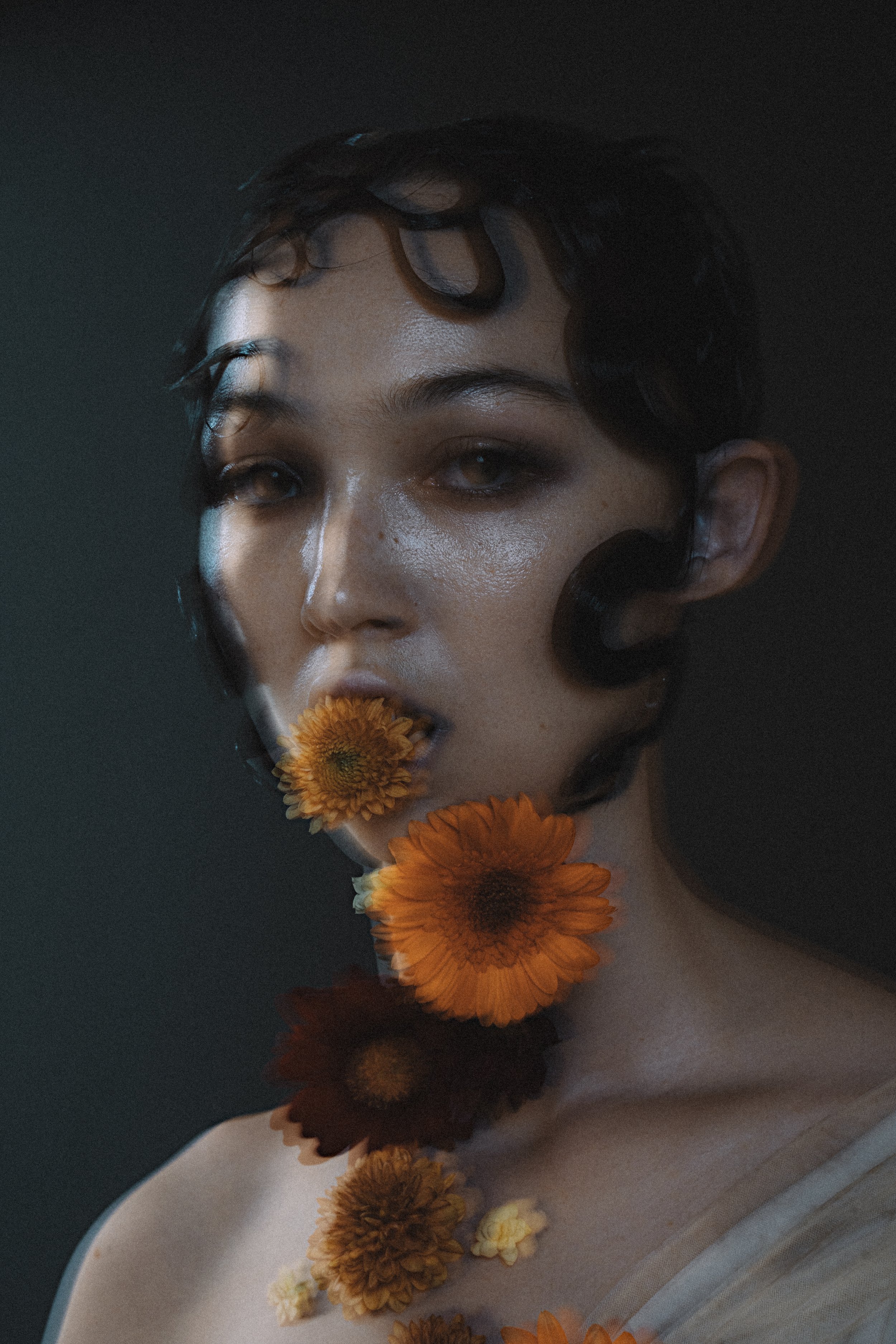 Portrait of a woman with short dark hair holding orange and yellow flowers in her mouth and on her neck against a dark background.