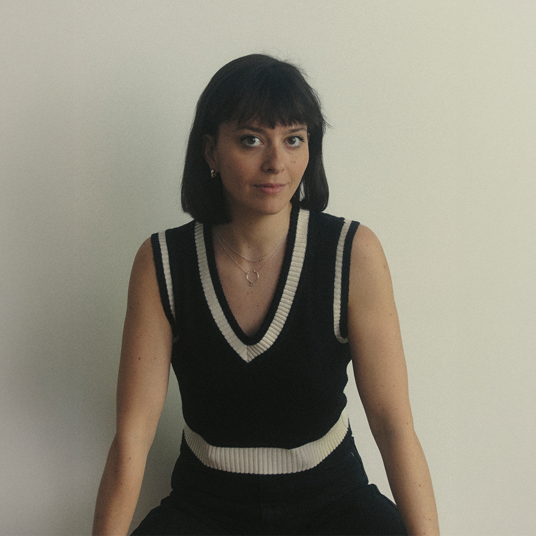 A woman with short dark hair wearing a black sleeveless sweater with white and gray stripes, sitting against a plain white wall.