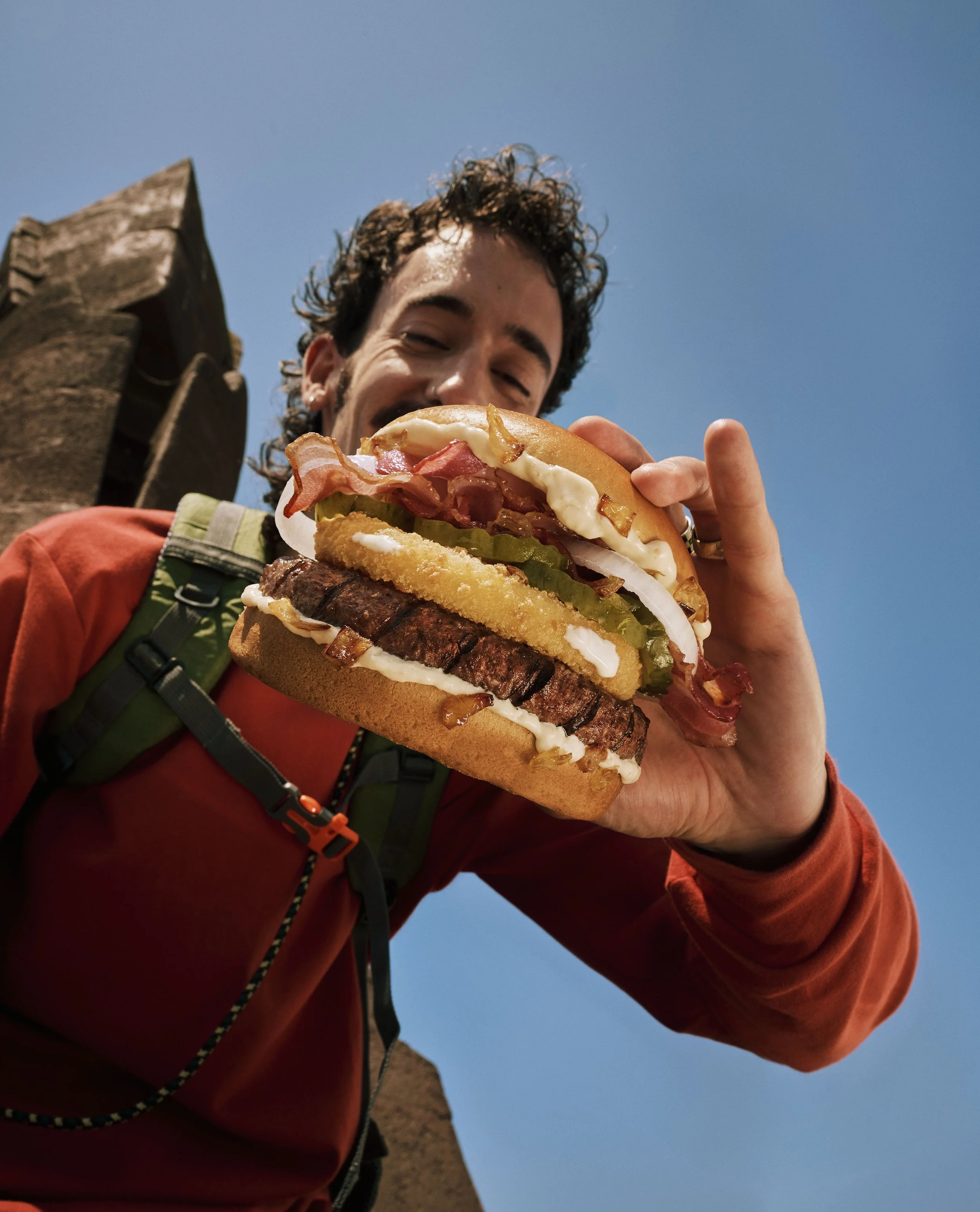 Person holding a large sandwich with layers of bacon, lettuce, tomato, fried onion ring, and condiments against a blue sky.