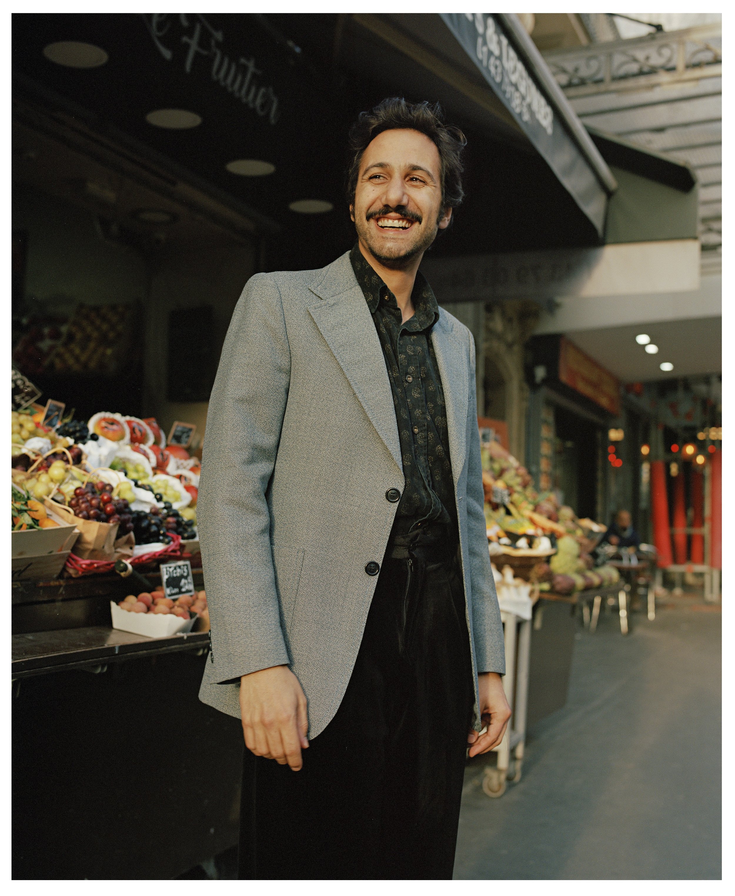 A man with dark hair and a beard smiling while standing in front of a fruit stand at an outdoor market. He is wearing a light gray blazer over a dark patterned shirt.