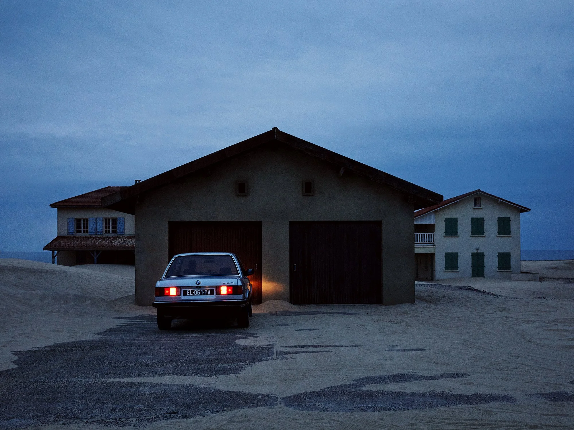 A parked car in front of a large garage on a sandy lot at dusk, with two houses in the background under a cloudy sky.