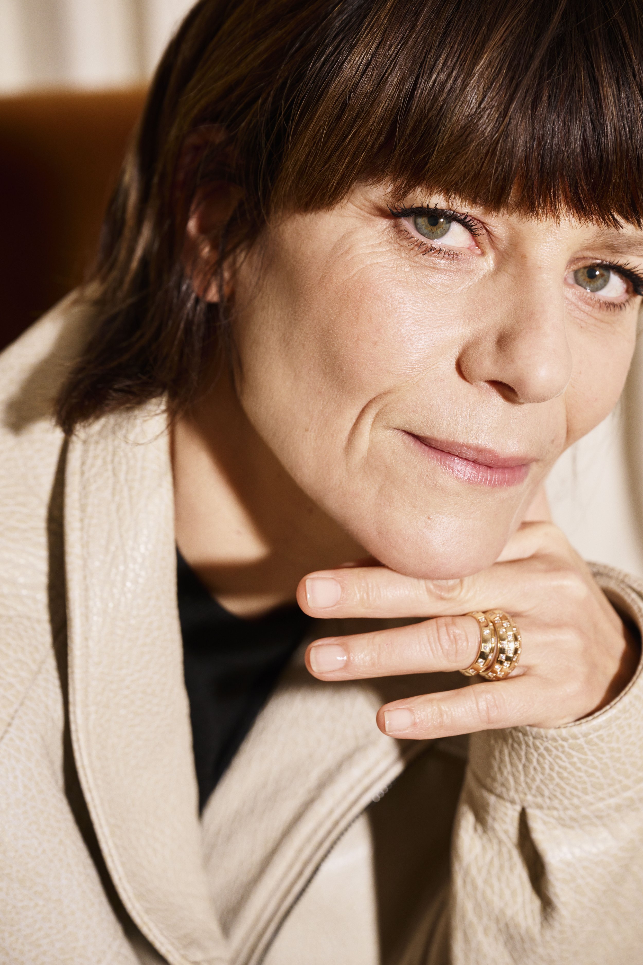 Close-up portrait of a confident woman with short brown hair, green eyes, wearing subtle makeup, a beige textured blazer, and multiple rings on her finger, resting her chin on her hand.