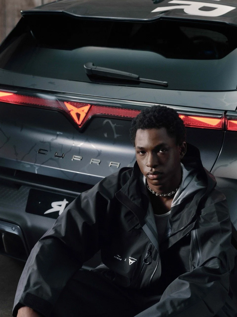 A young man with dark curly hair wearing a black jacket and a silver chain, sitting in front of a sleek grey CUPRA car with illuminated red rear lights.