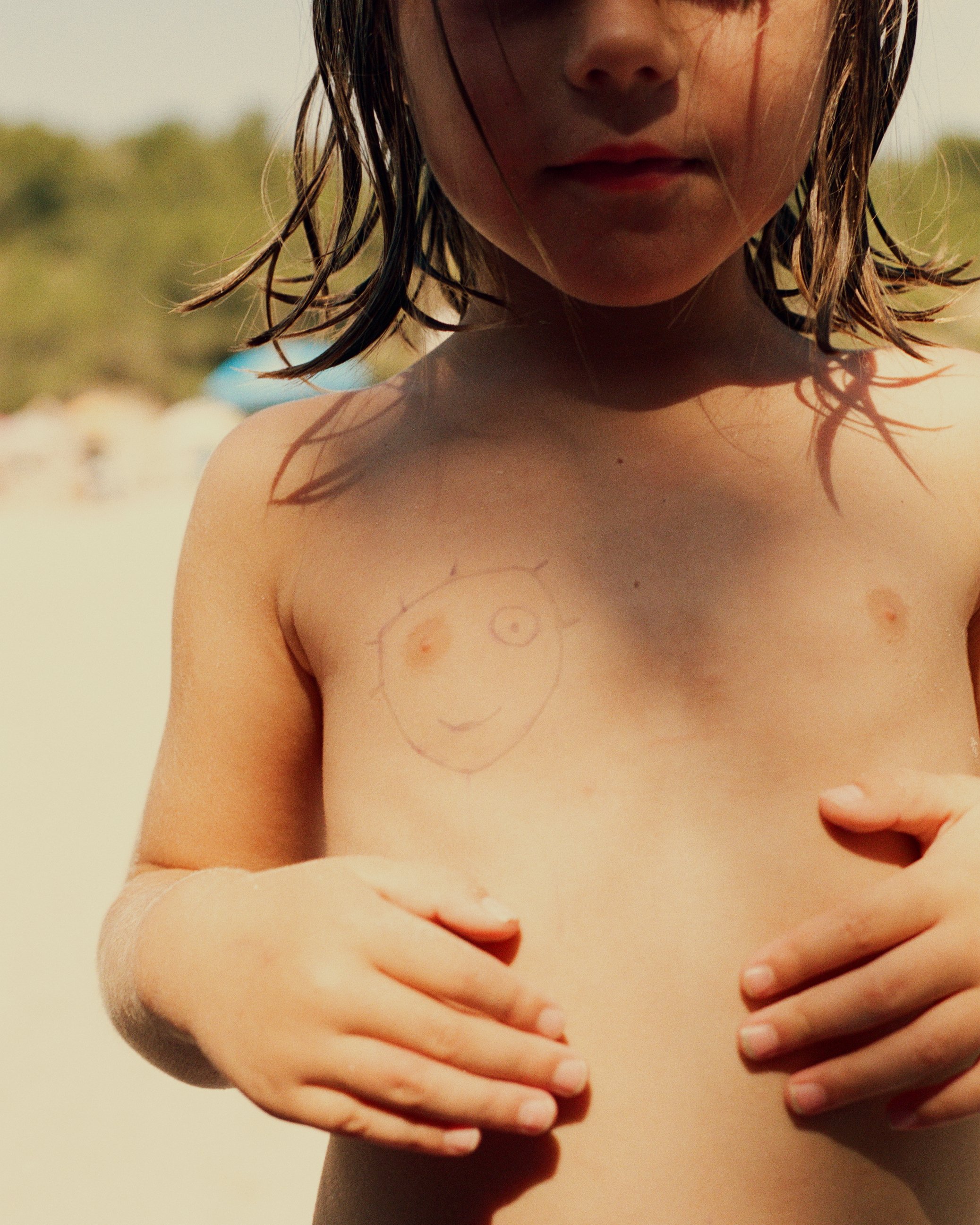A young girl at the beach with wet hair, holding her breasts with both hands. There is a child's drawing of a smiling face with one eye on her chest.