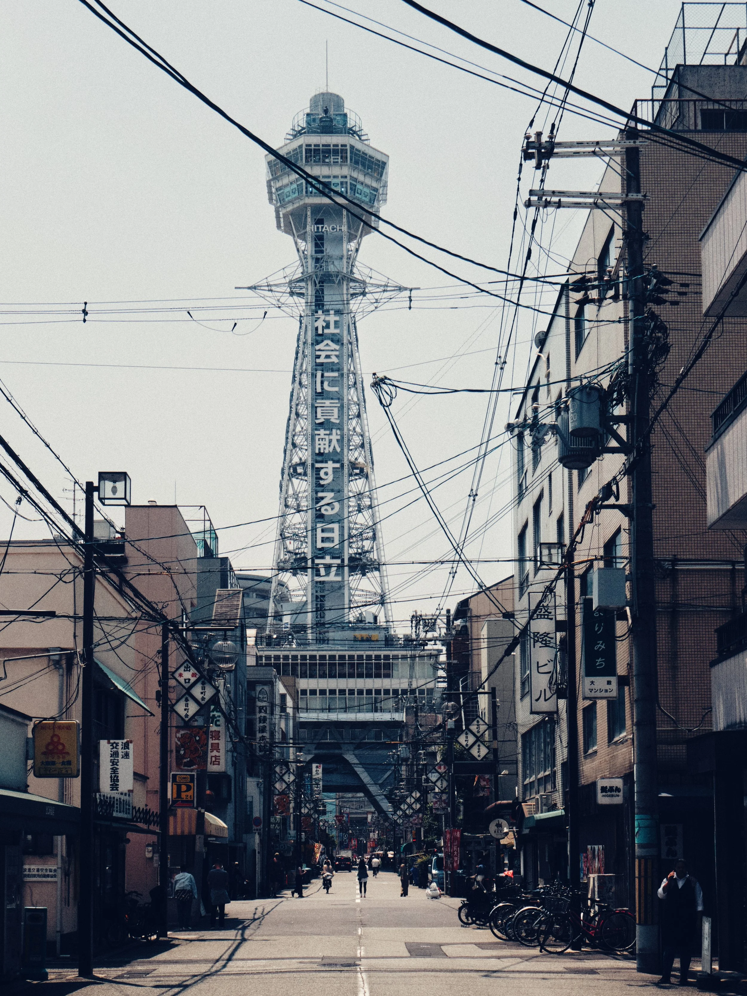 An urban street scene in Japan with the Tokyo Skytree in the background, surrounded by power lines and buildings, with pedestrians and bicycles on the street.