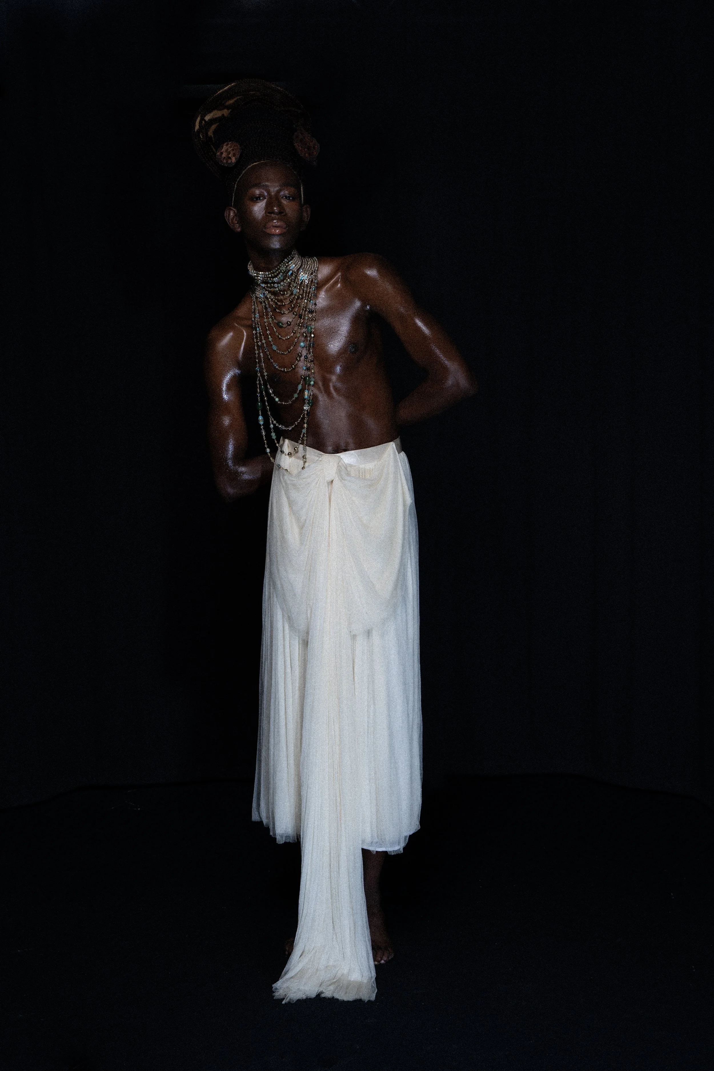 A person with dark skin tone, wearing multiple layered jewelry, and dressed in flowing white trousers, standing against a black background.