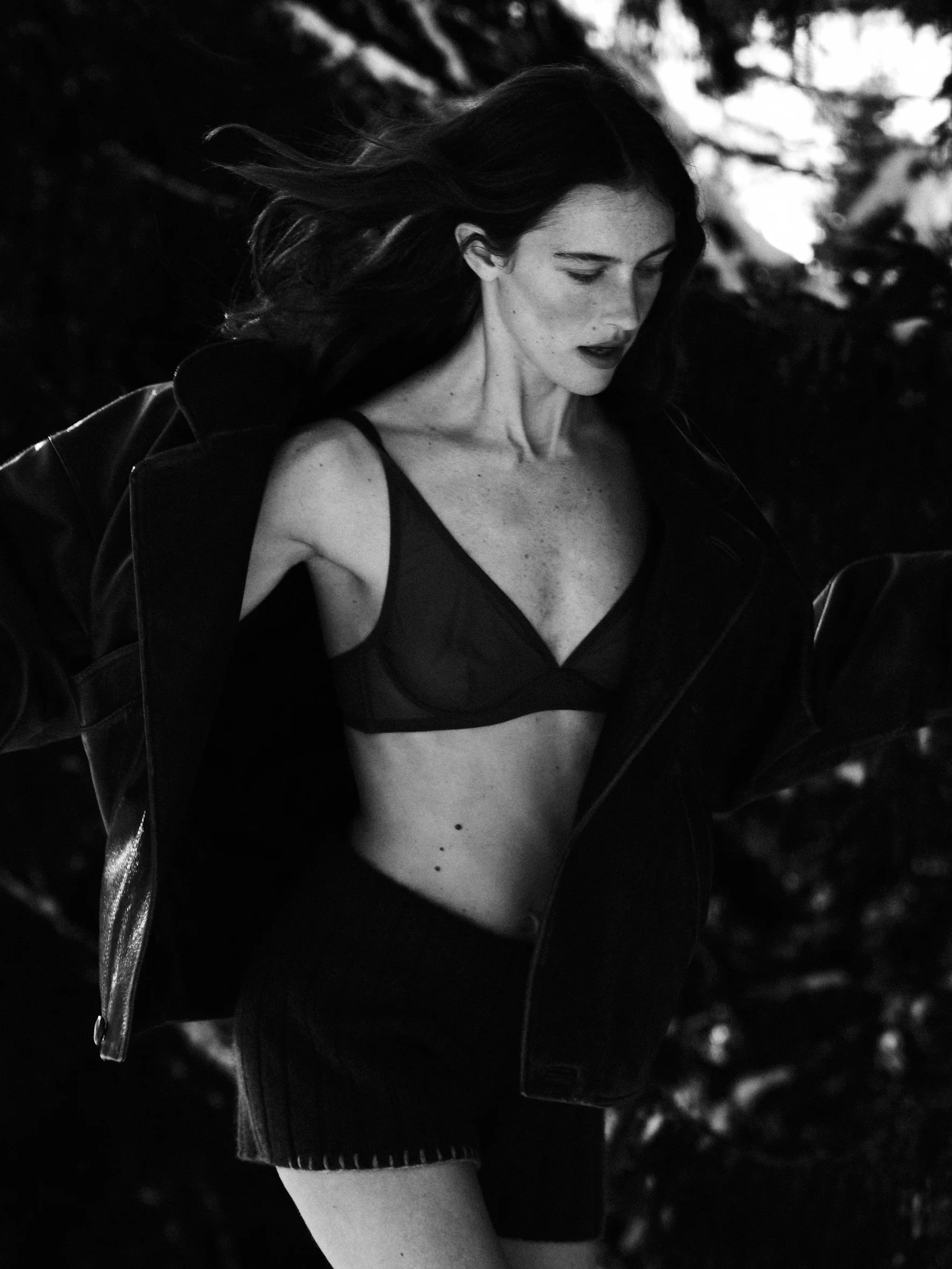 A black and white photo of a woman with long hair, wearing a bra and shorts, outdoors with blurred trees in the background.