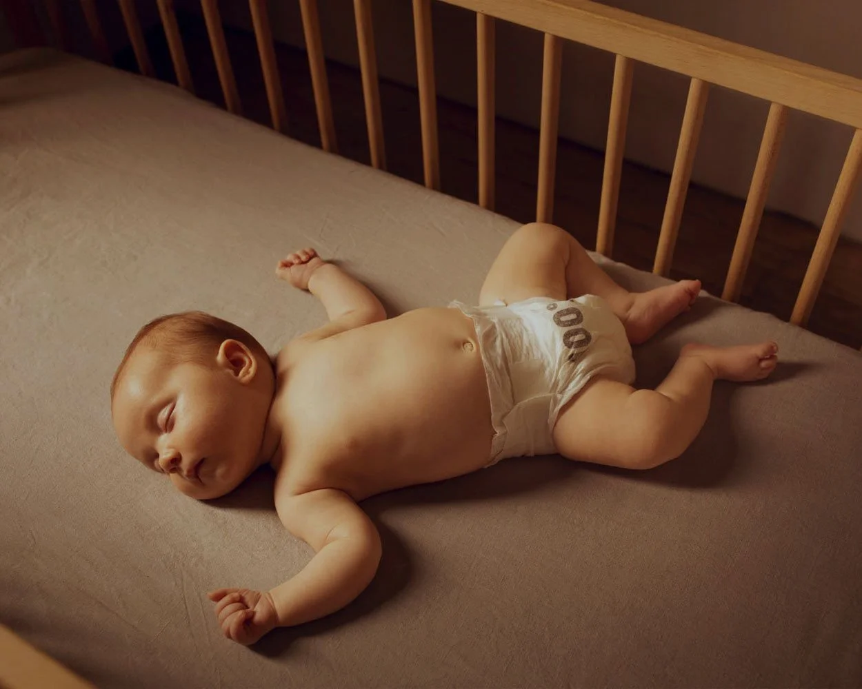 A baby sleeping on a bed, wearing only a diaper, with arms outstretched and eyes closed.