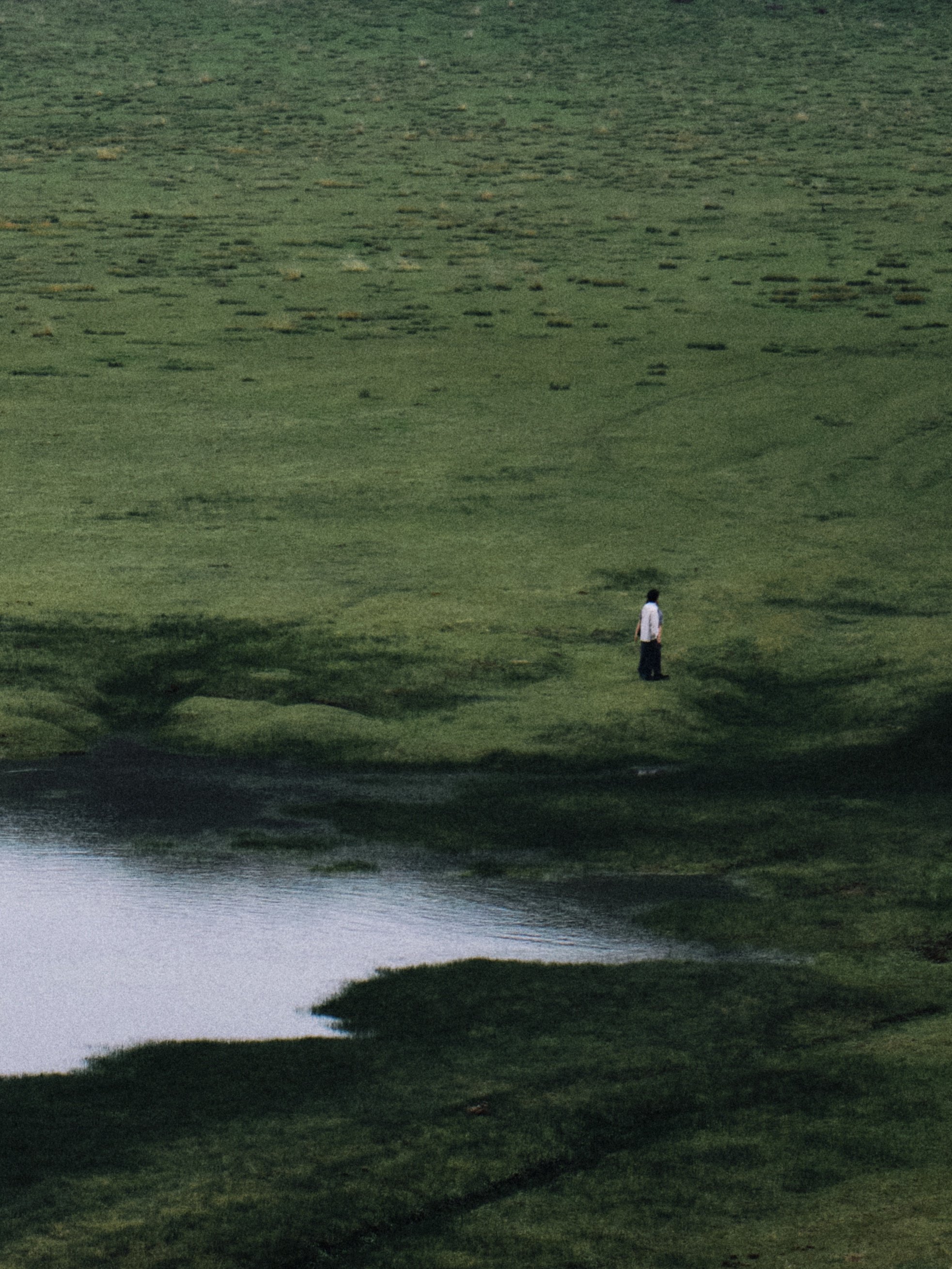 A person standing on a grassy land near a small body of water, with lush green landscape in the background.