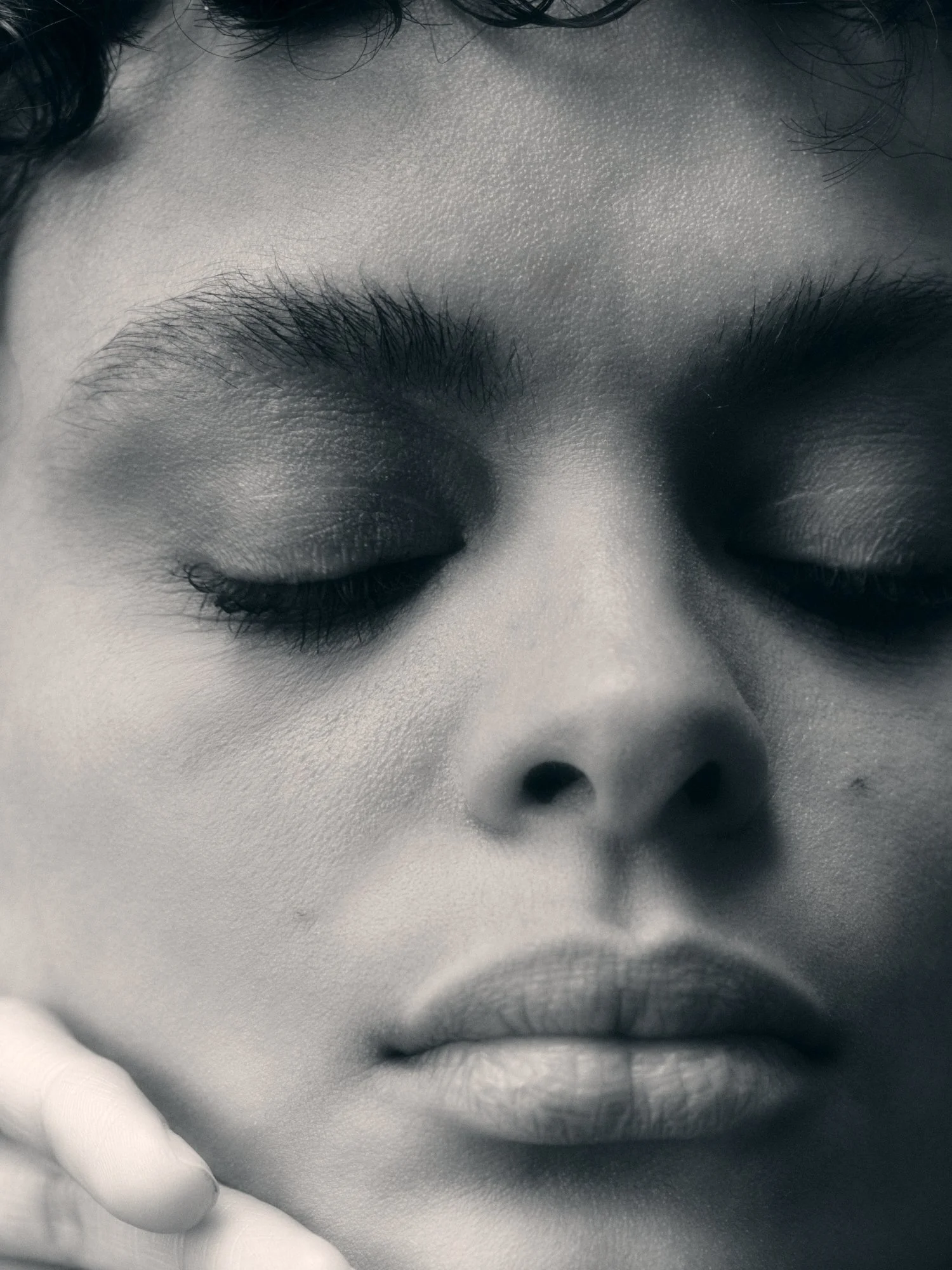 Close-up of a person's face with closed eyes, showing detailed skin texture on the forehead, eyebrows, and nose, in black and white.