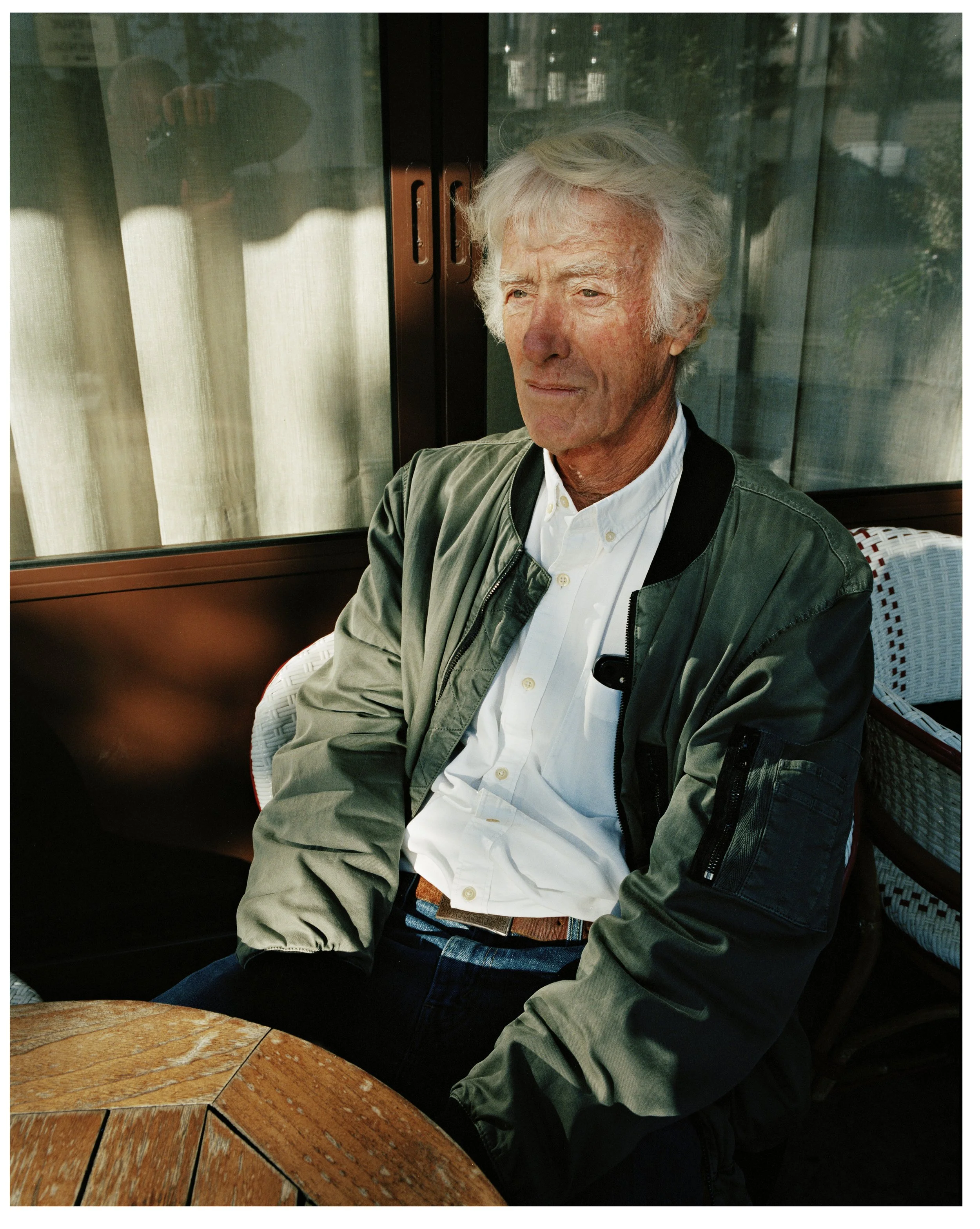 An older man with white hair sitting at an outdoor table on a patio, wearing a white button-up shirt and a green bomber jacket, looking thoughtful.
