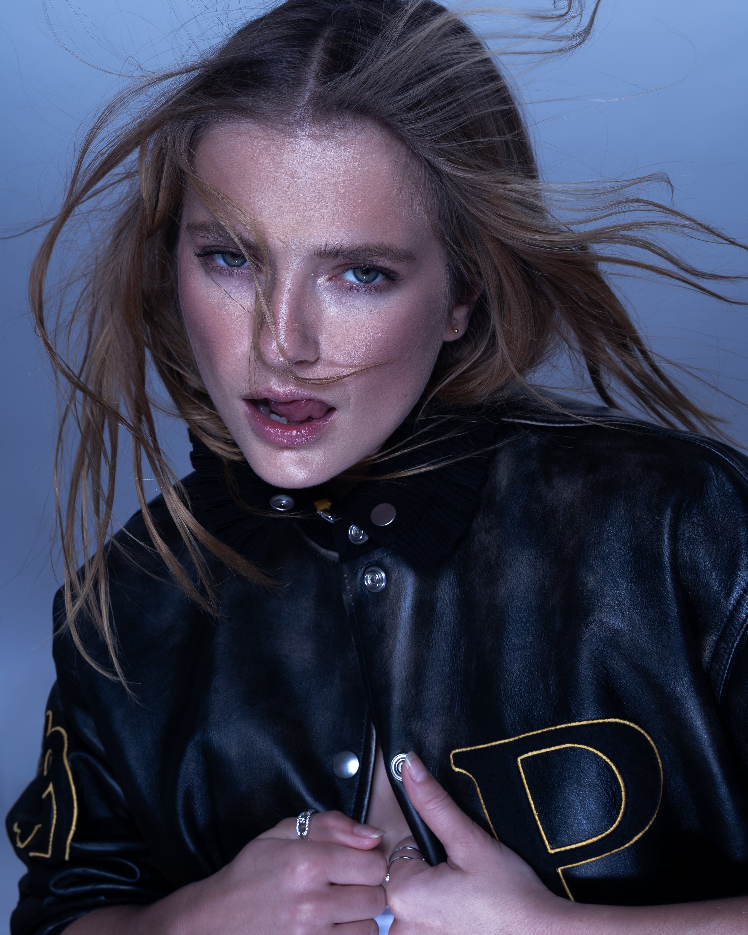 A woman with blonde hair and blue eyes, wearing a black leather jacket, sticking her tongue out and making a playful expression.