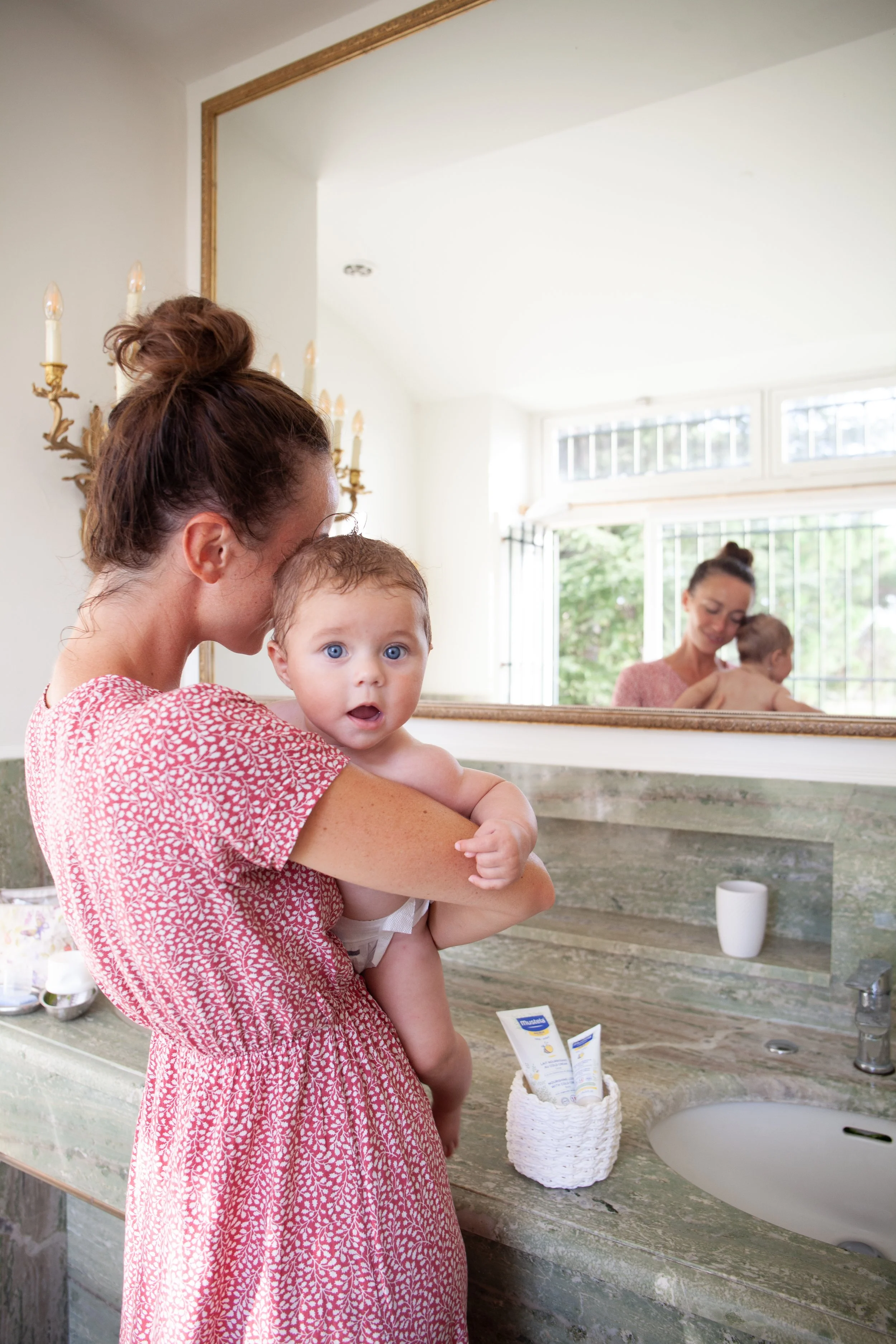 A woman holding a baby in a bathroom, looking into a mirror. The woman is wearing a pink patterned dress, and the baby is looking directly at the camera with wide eyes. The bathroom has a marble countertop, a woven container with toiletries, and a la