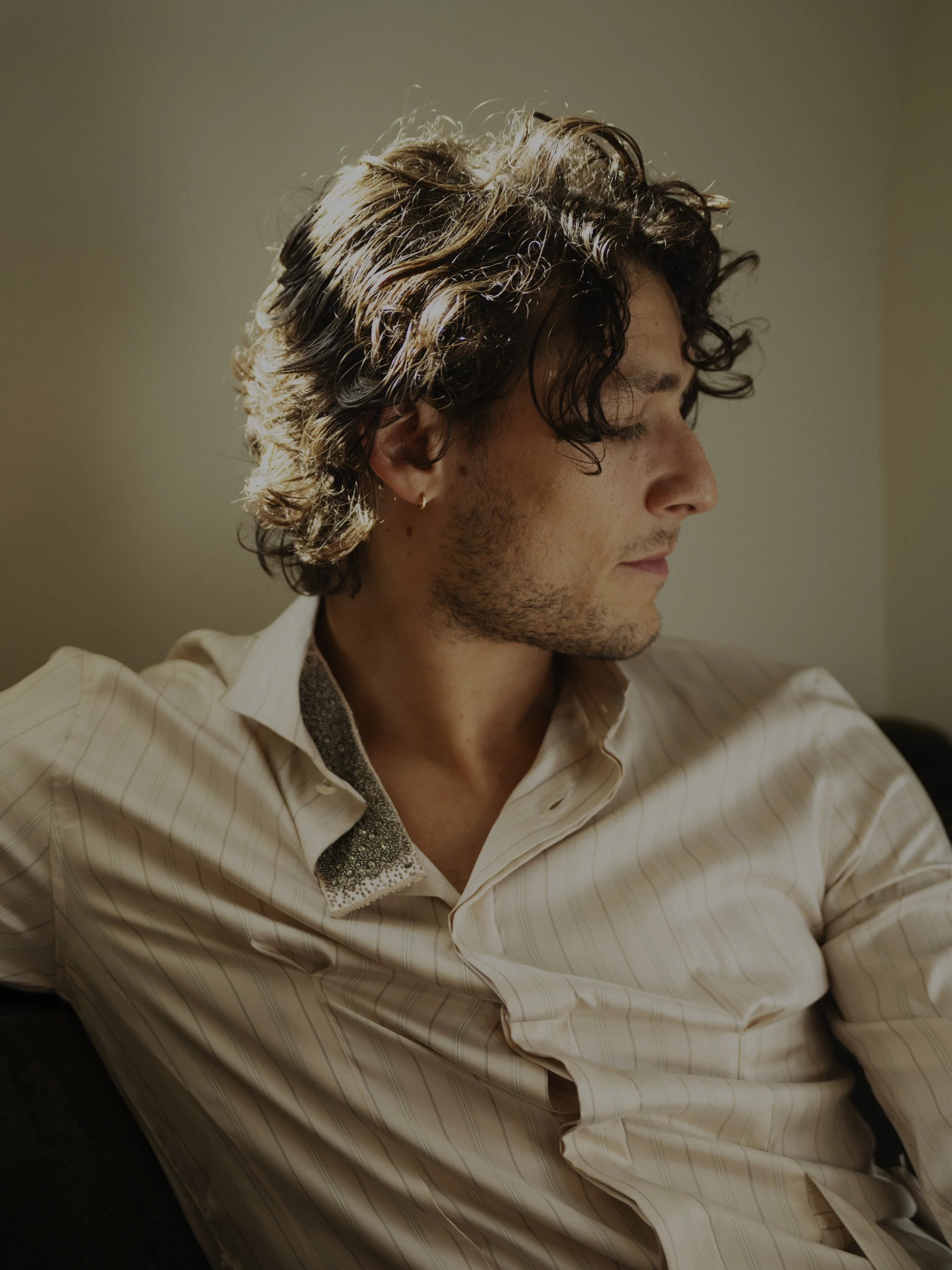 A man with dark curly hair and a earring, wearing a beige striped shirt, is looking to the side with a calm expression.