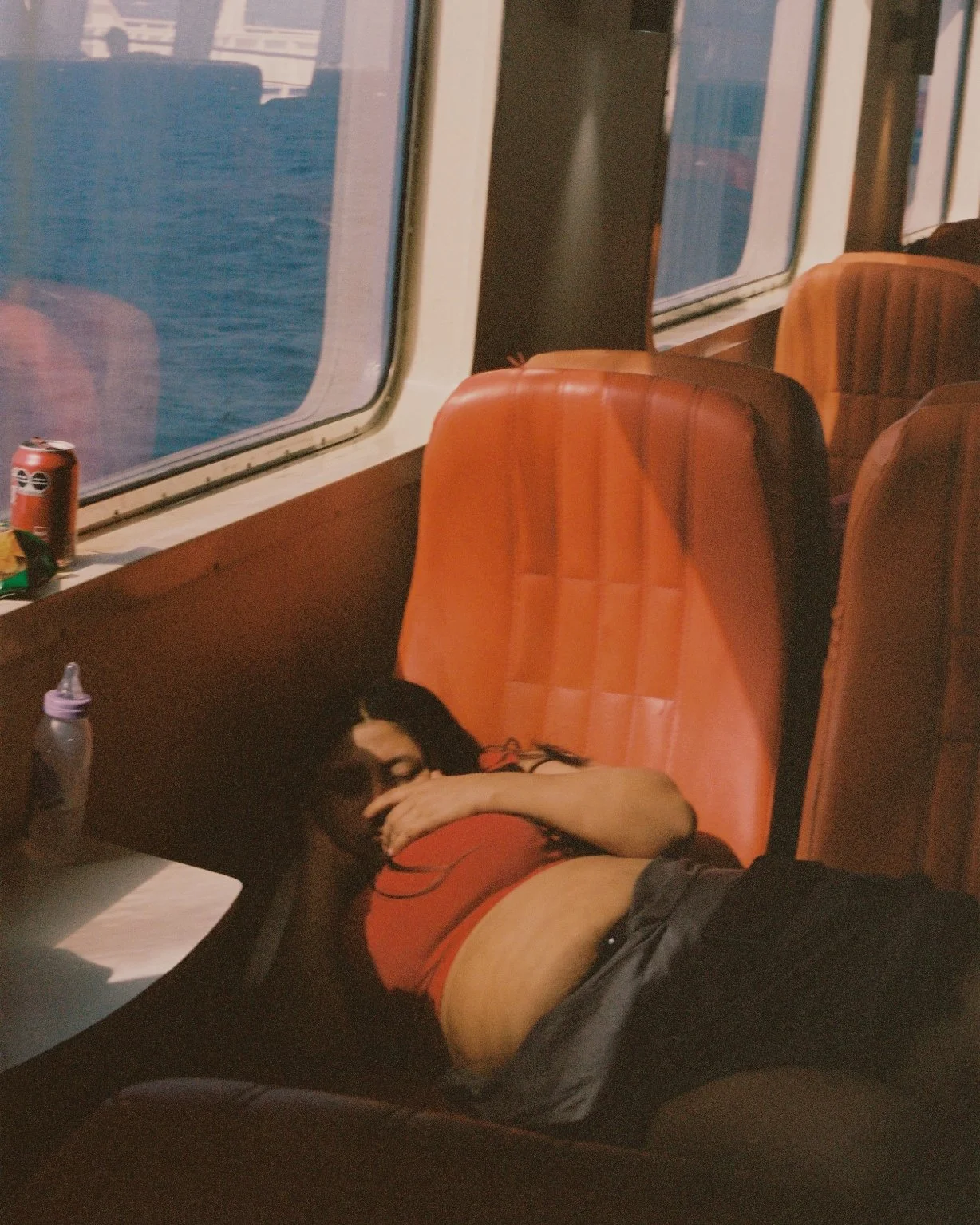 A woman with dark hair is sleeping on a bus seat, lying on her side with her arm over her face. She is wearing a red top and black shorts. There are orange bus seats, a window showing water outside, and a windowsill with a can and some items.