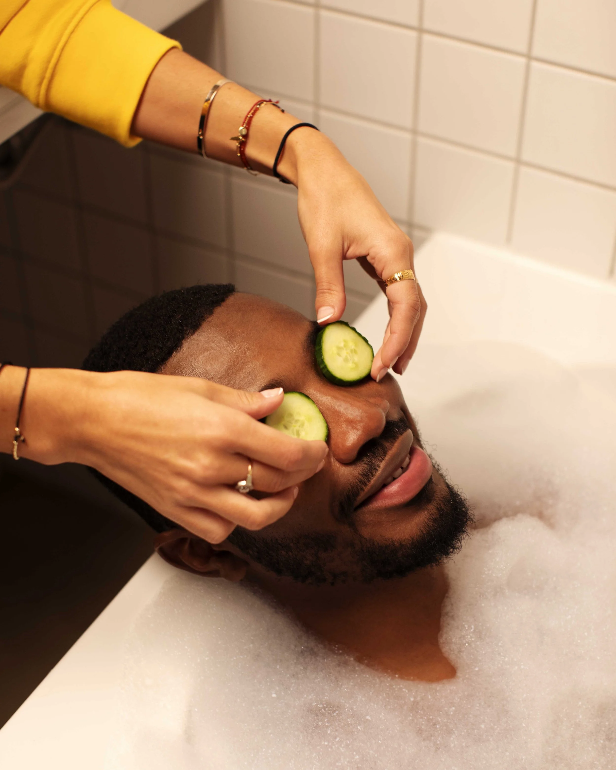A man relaxing with cucumber slices on his closed eyes while a woman gives him a facial treatment in a bubble bath.