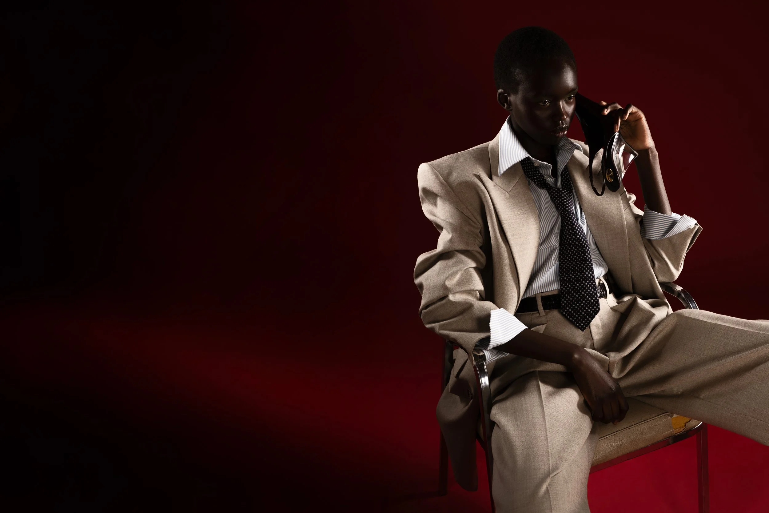 A person in a beige suit, white shirt, and black tie sitting on a chair, holding a telephone to their ear against a dark red background.