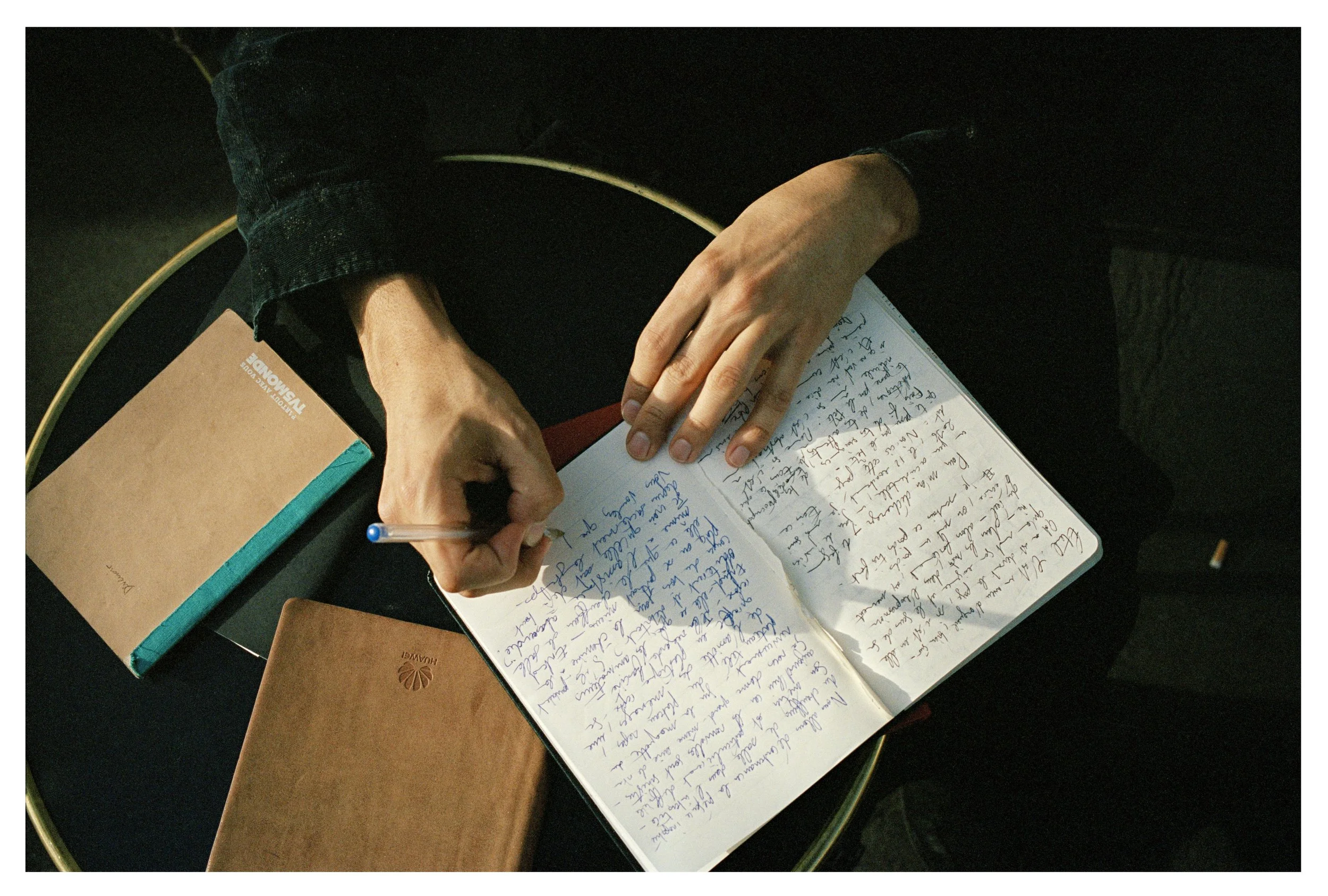 Person writing in a notebook with blue ink, with two brown notebooks and a smartphone on a black table.