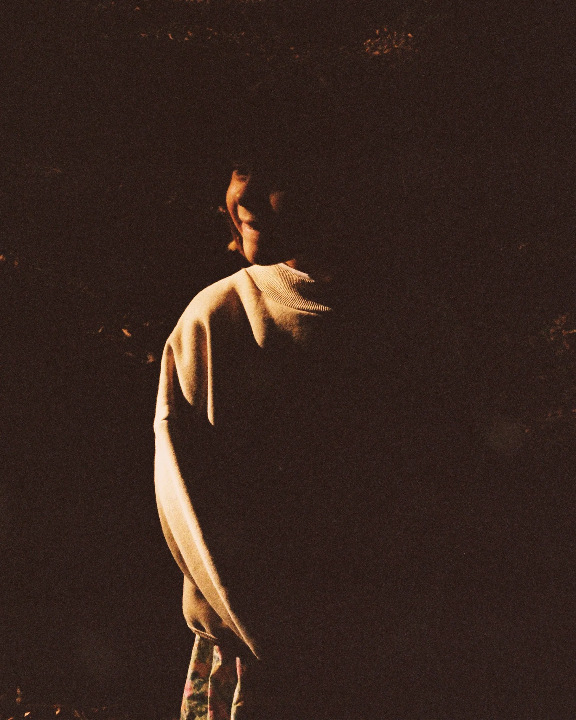 A person standing in shadow with sunlight illuminating part of their face and clothing, against a dark background.