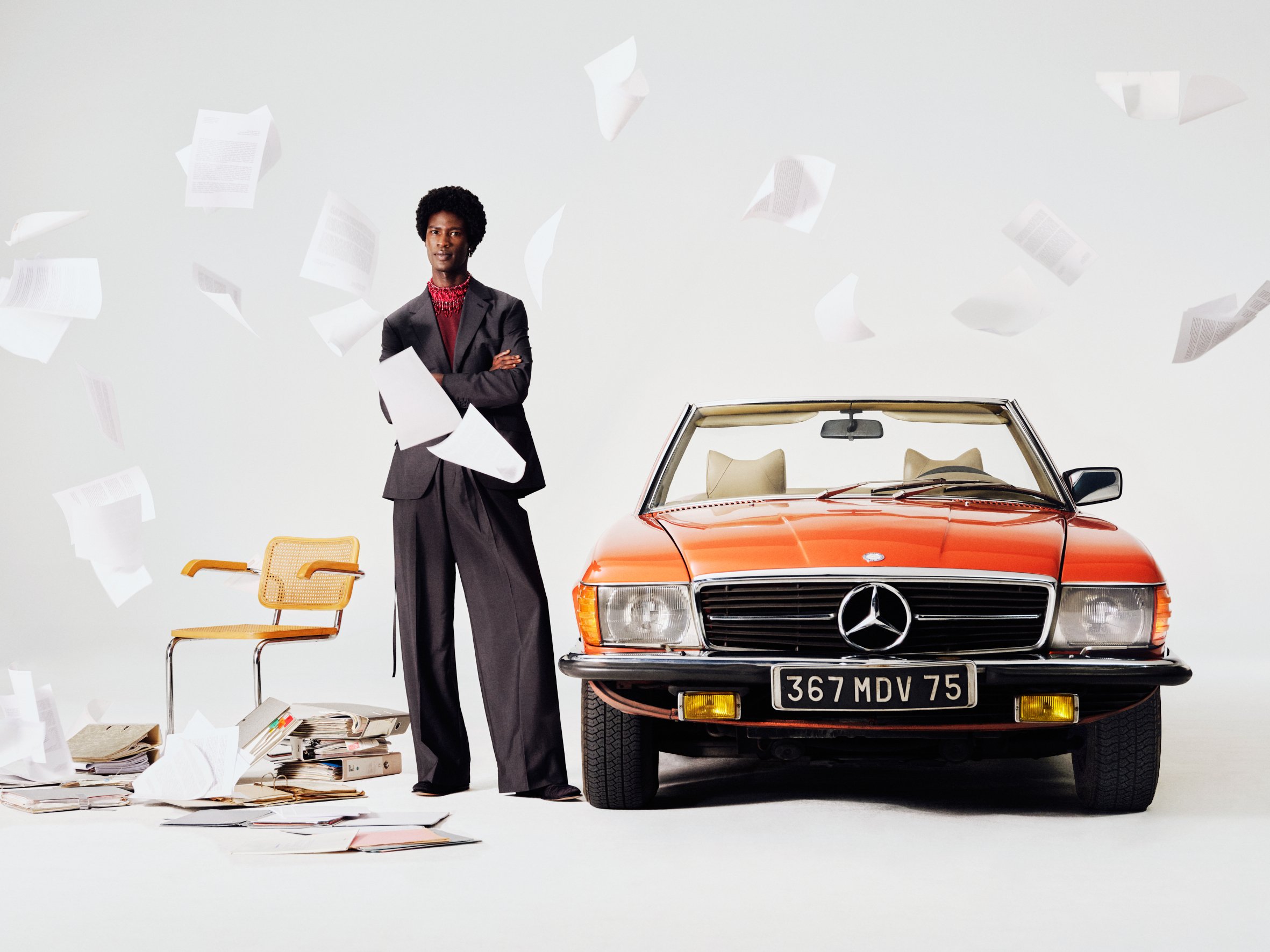 A woman in a business suit standing with arms crossed next to a vintage orange Mercedes-Benz convertible car, with papers flying around and scattered on the floor.