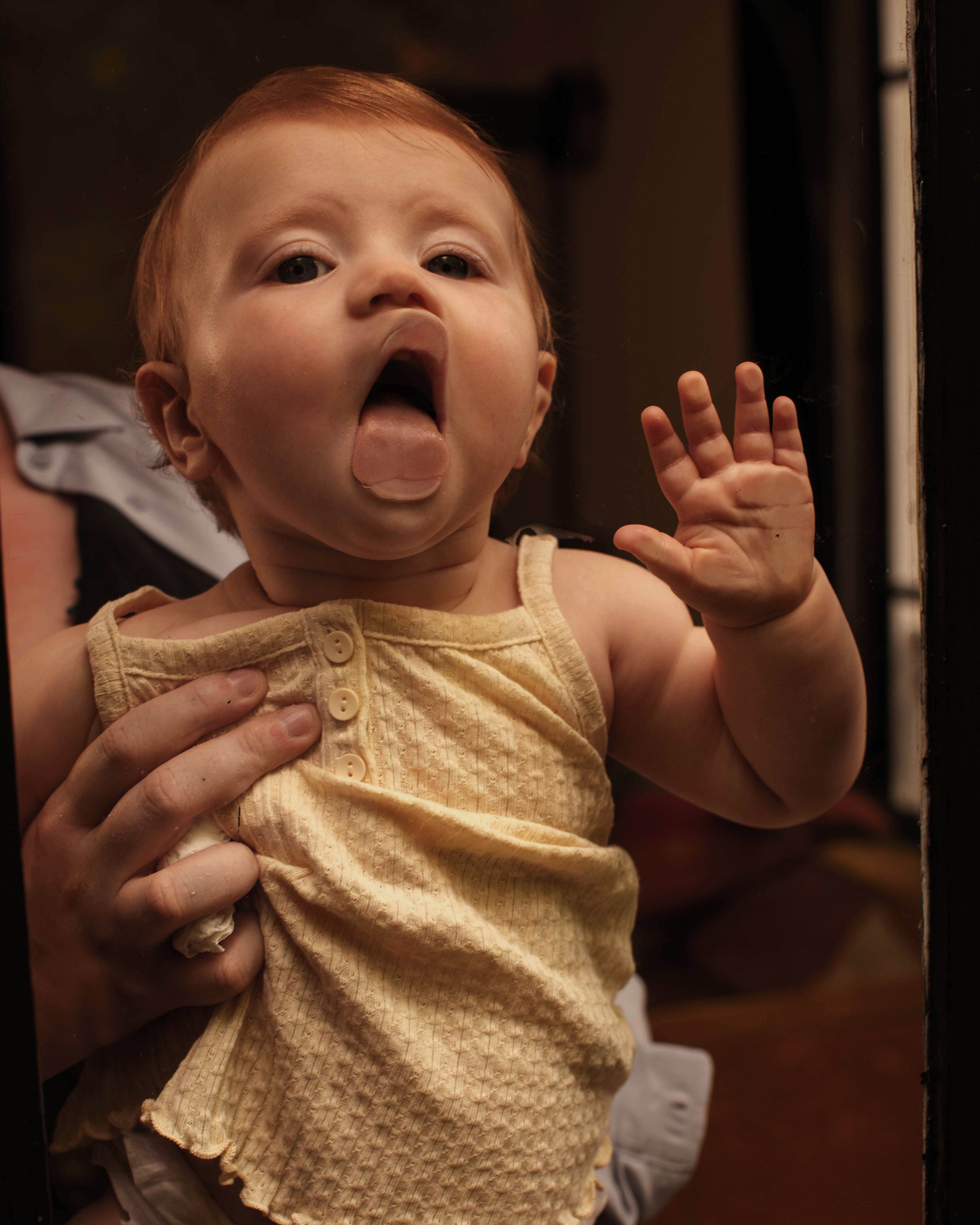 A young child with red hair sticking out their tongue and raising one hand as someone holds them from behind.