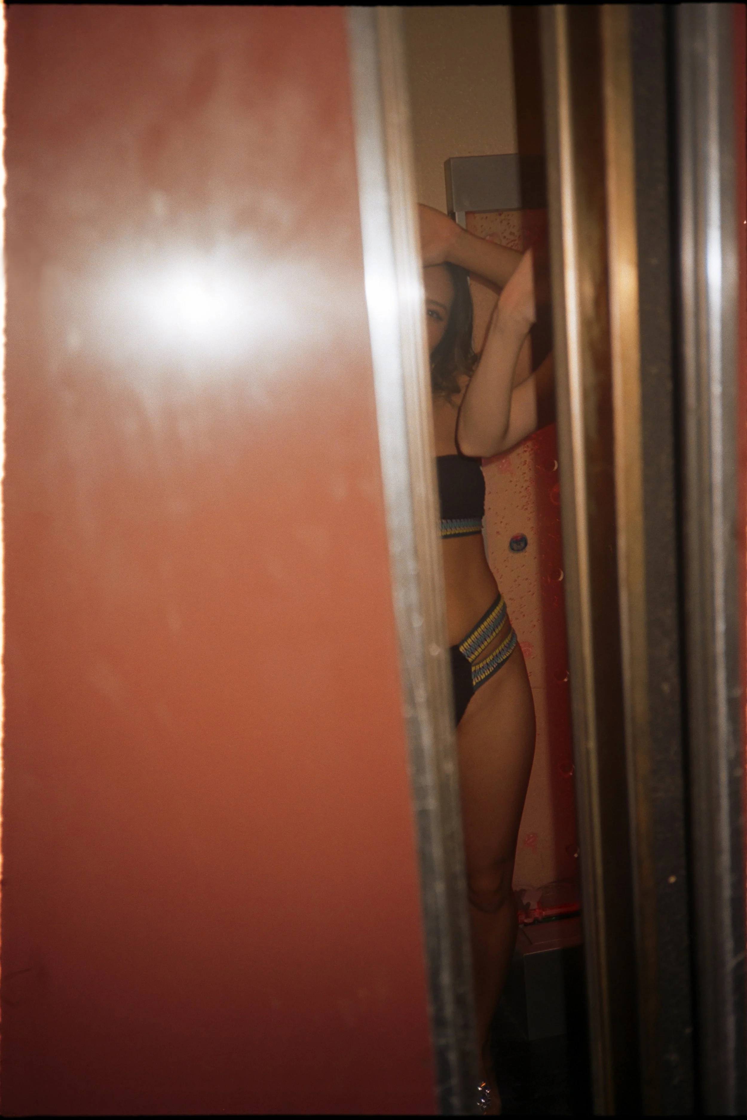 A woman in a black top and colorful patterned underwear is taking a mirror selfie in a changing room, with her face partially visible behind the door.